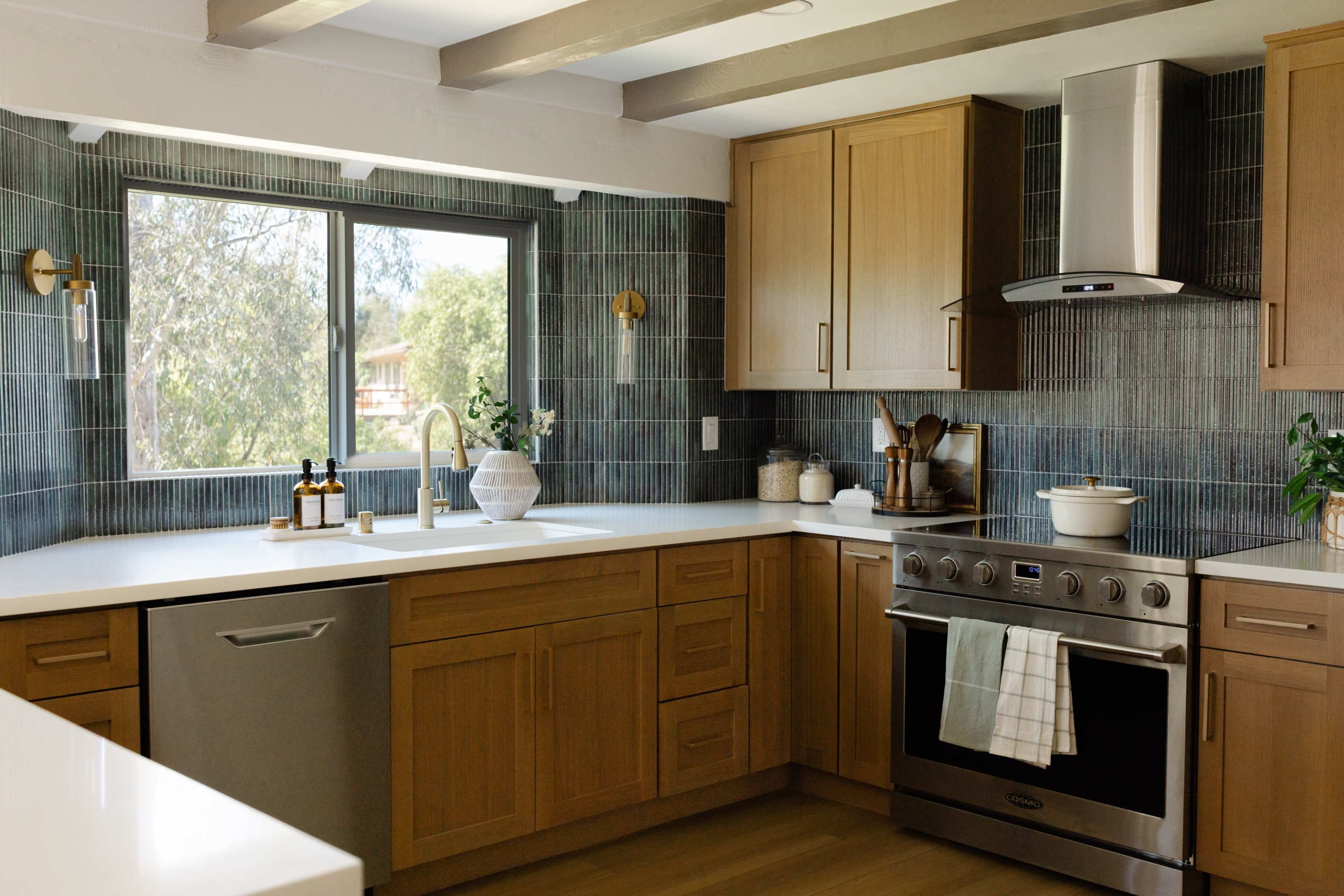 The image shows a modern kitchen with wooden cabinets, a stainless steel stove, and a large window allowing natural light to enter.