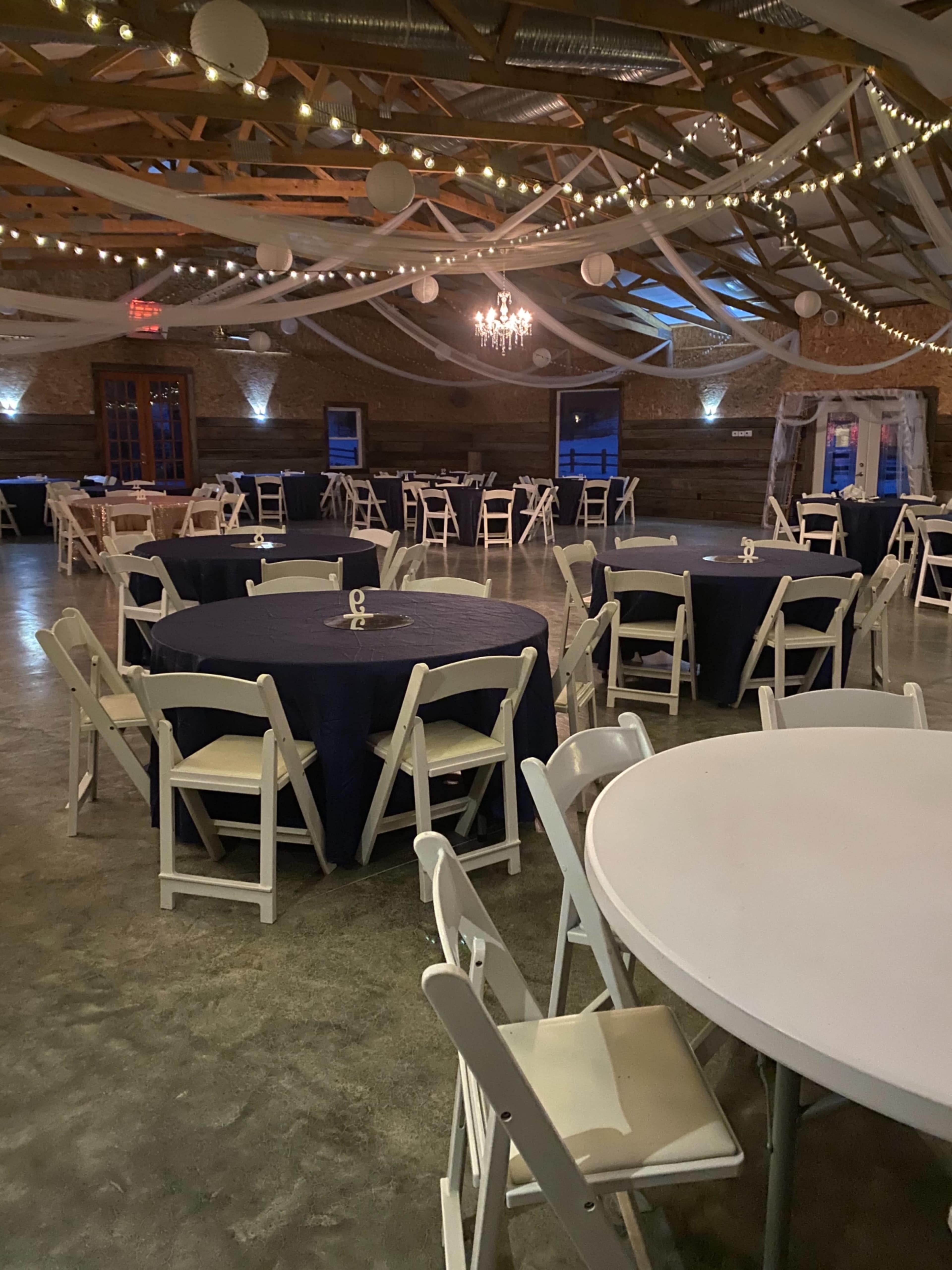 The image shows an indoor event space with round tables covered in dark blue tablecloths and decorated with white chairs, all beneath a canopy of lights and hanging decorations.