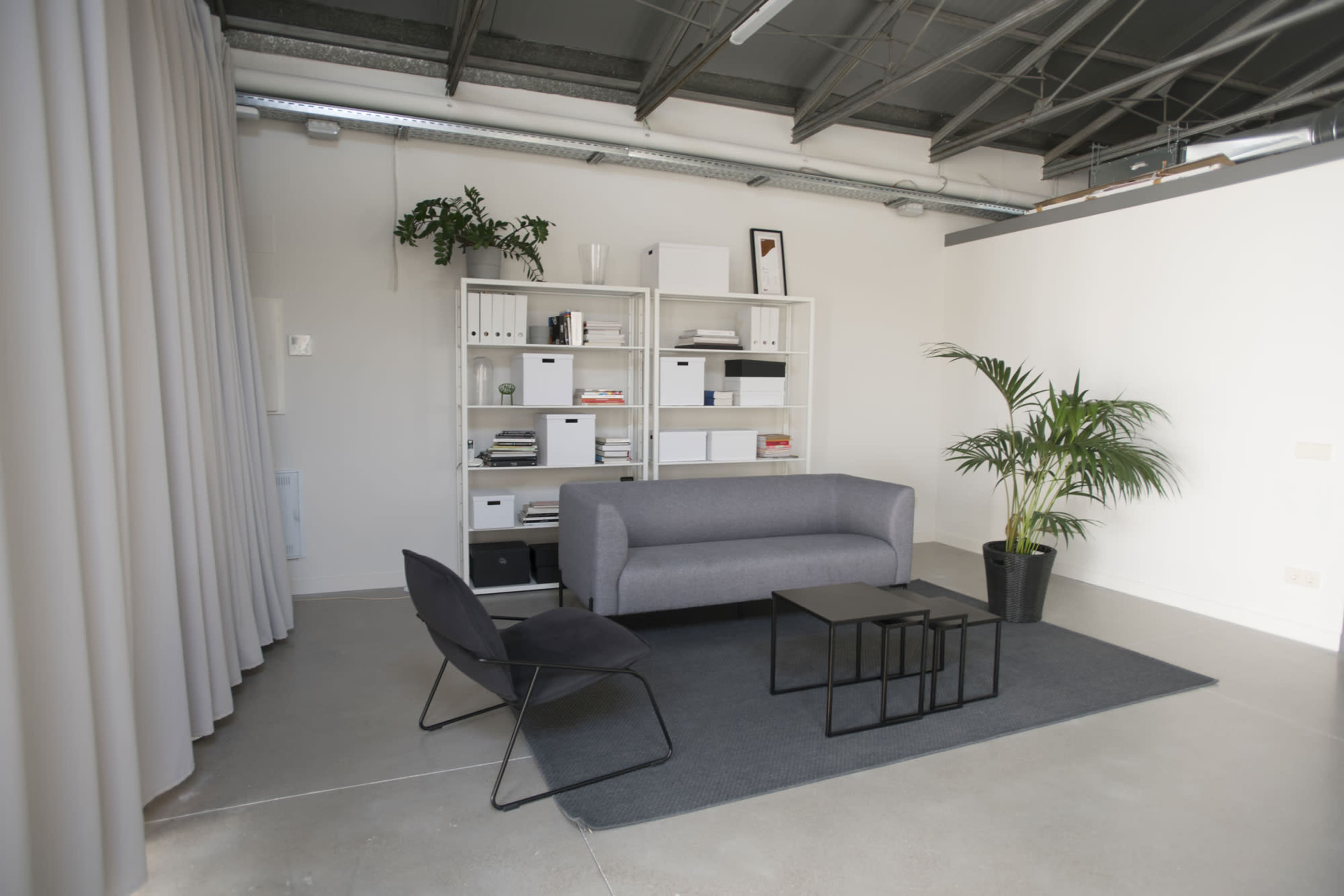 A minimalist office space features a gray couch, a black chair, a black coffee table, a light gray rug, and a shelving unit with storage boxes against a white wall.