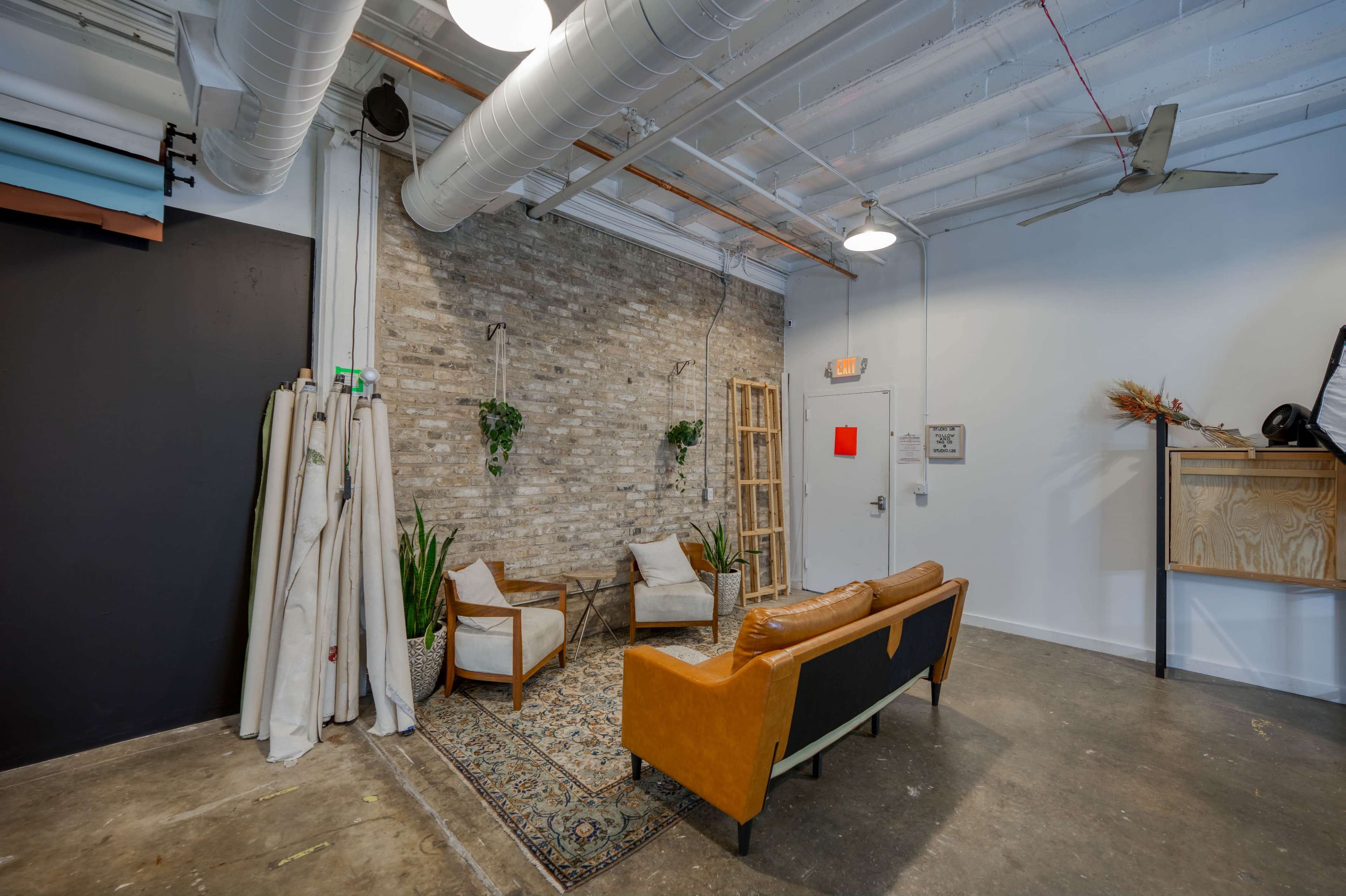 A modern interior space featuring a leather sofa, two chairs, and a patterned rug against a brick wall, with plants and a ladder in the background.