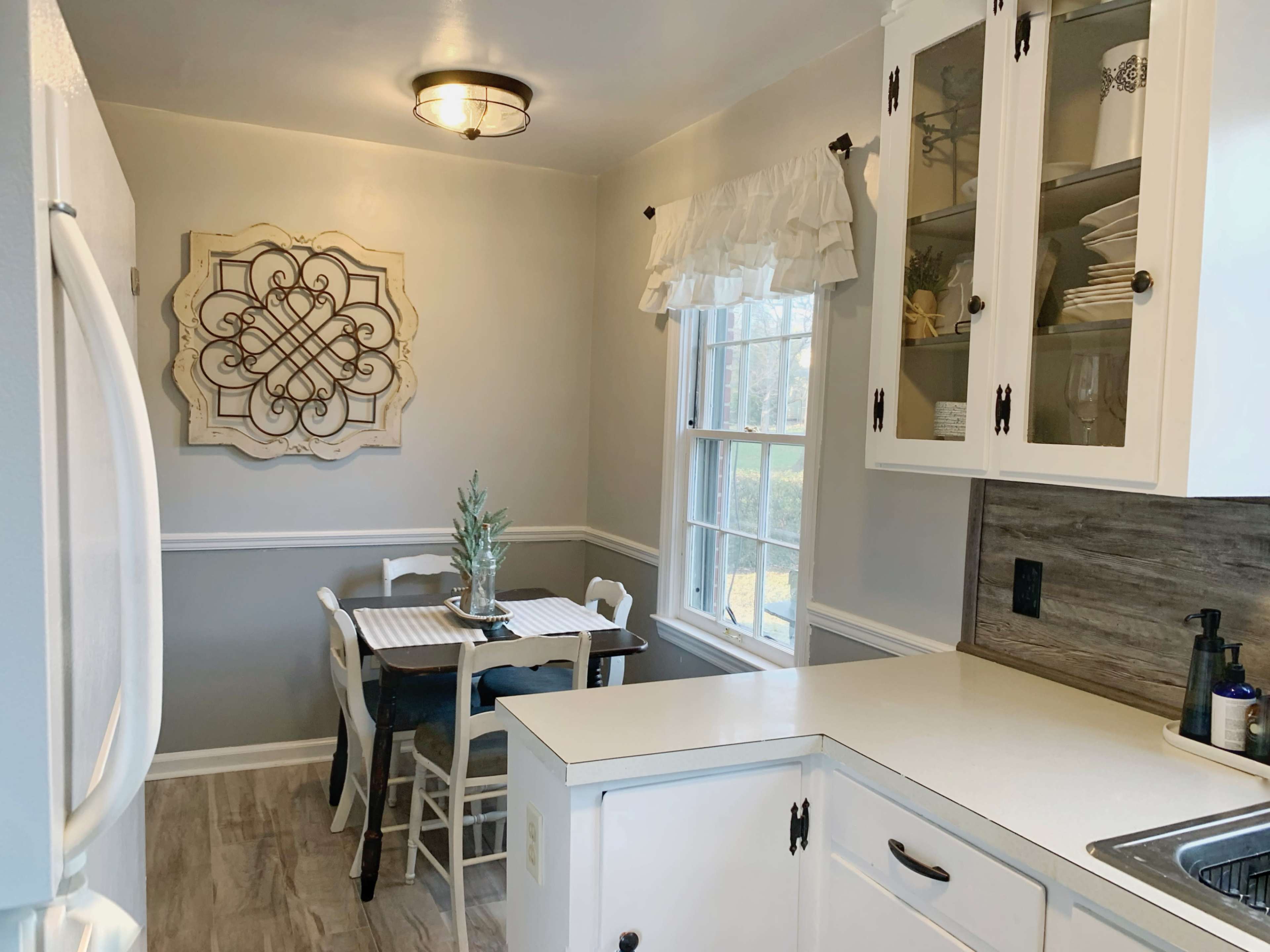 A bright and neatly organized kitchen features white cabinetry, a small dining table with chairs, and a decorative wall plaque.