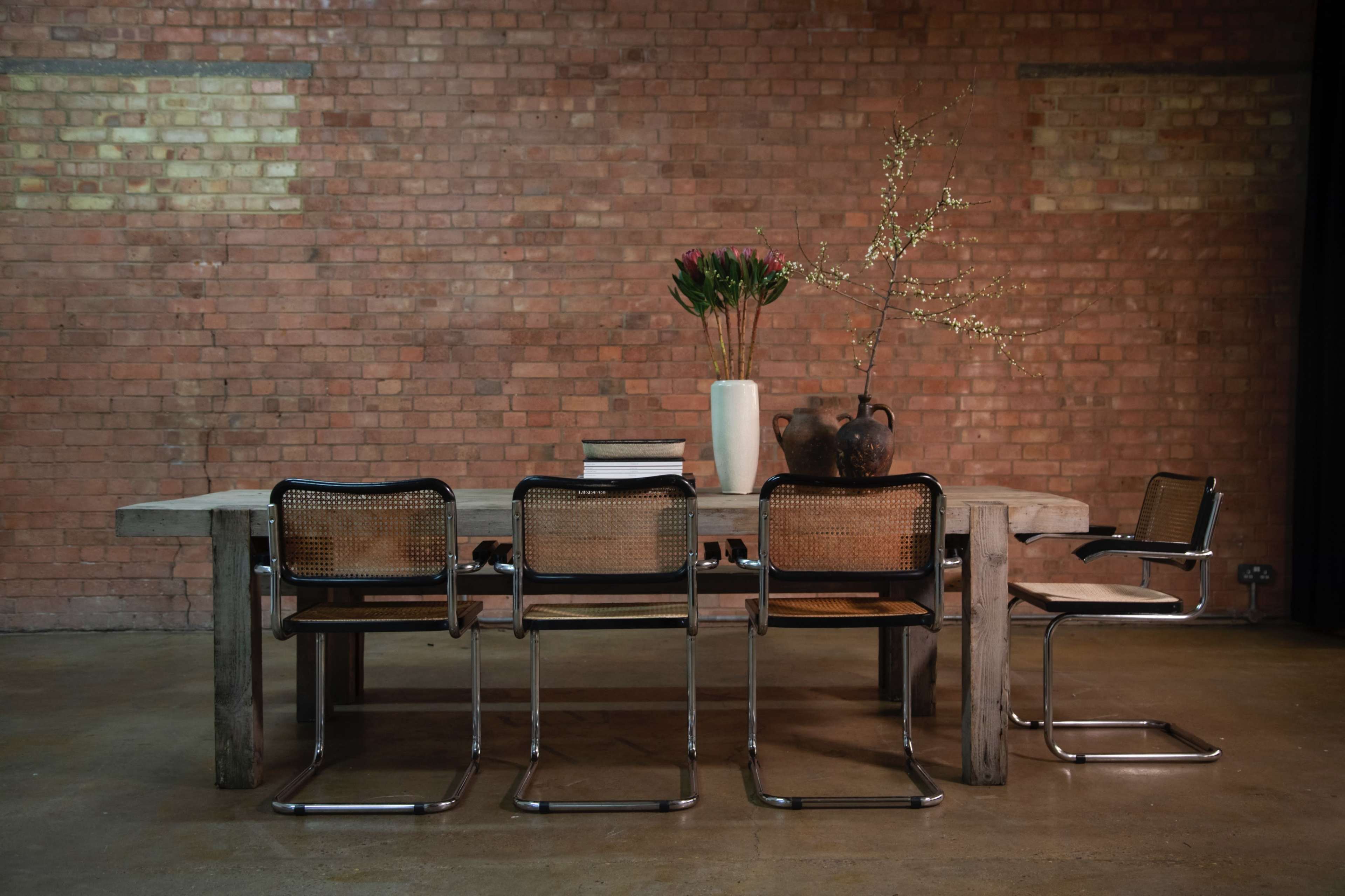 A long concrete table is set against a brick wall, accompanied by four metal and wood chairs, a vase with flowers, and a decorative pot.