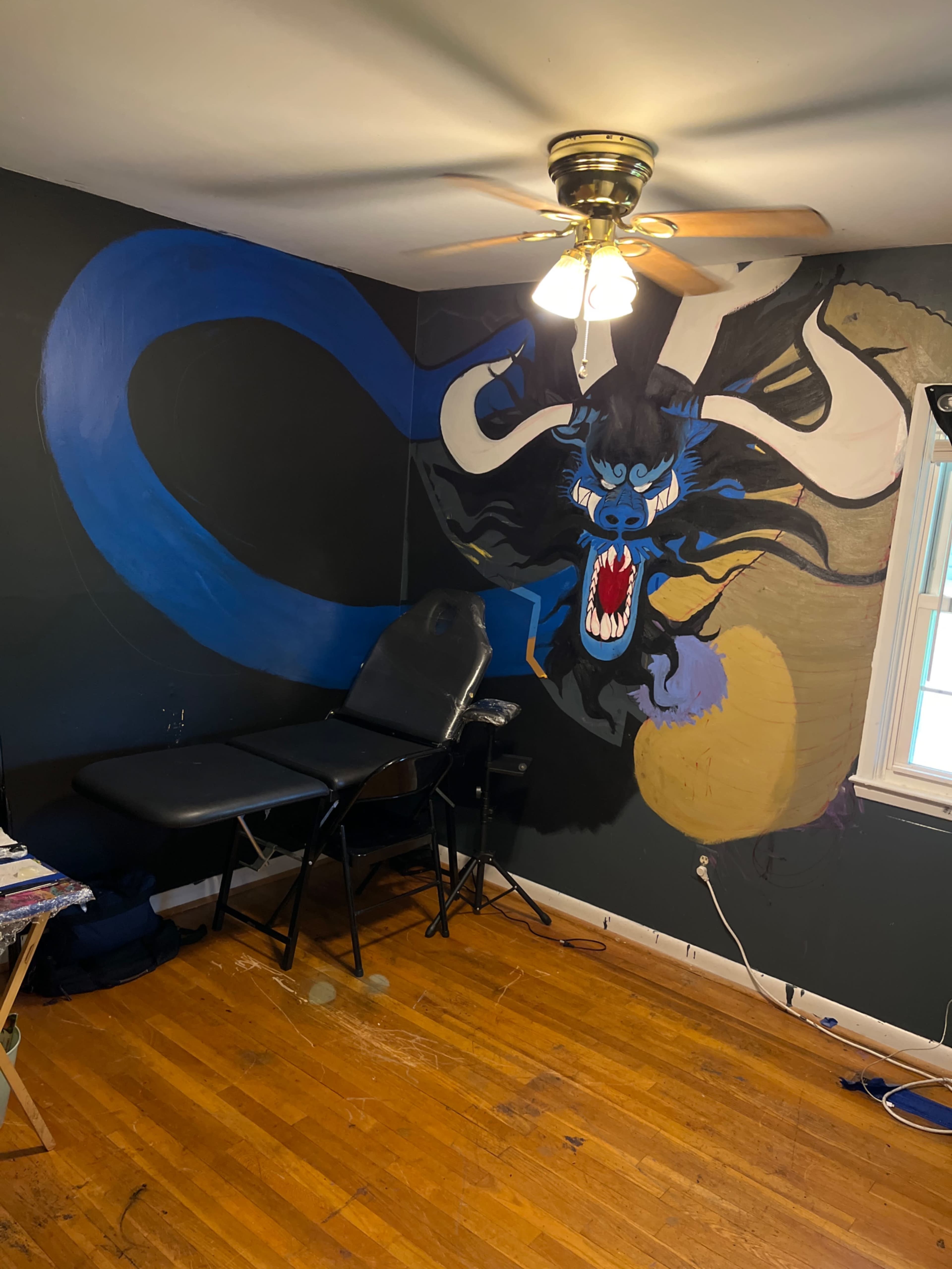 A room with hardwood flooring, featuring a tattoo chair and a large mural of a dragon on the wall.