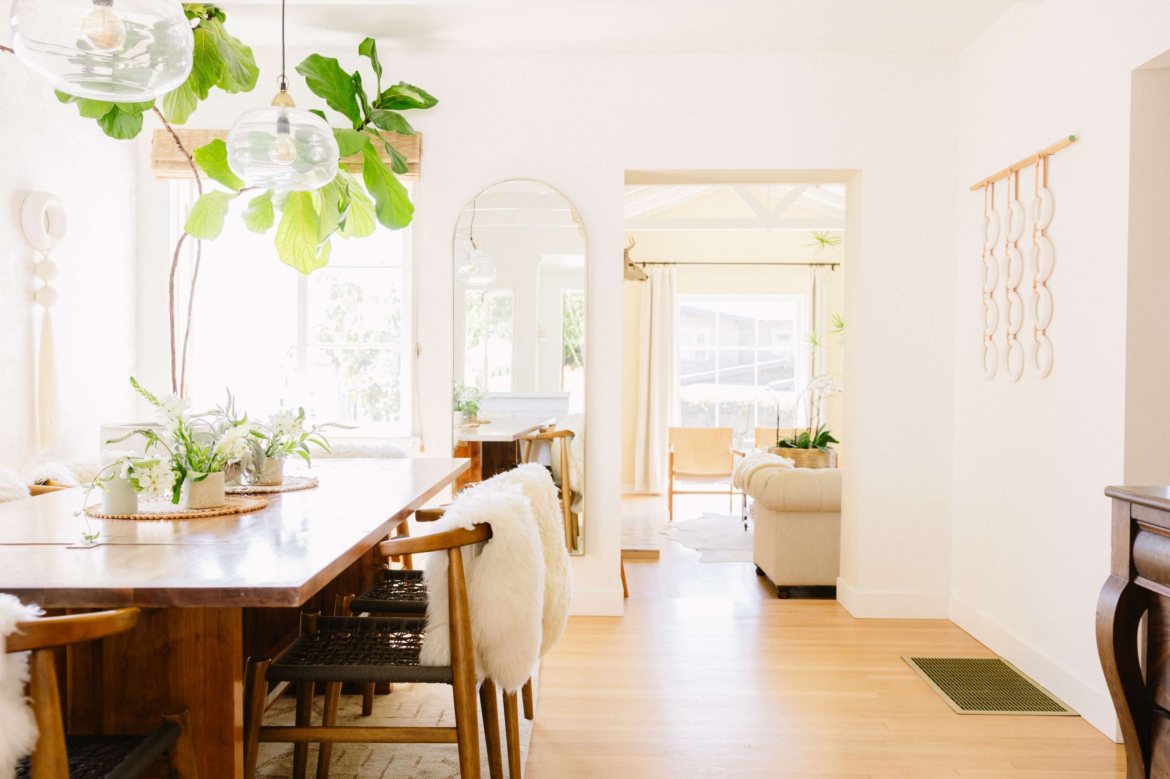 The image shows a bright, modern dining area with a wooden table, surrounded by plants, leading into a cozy living room.