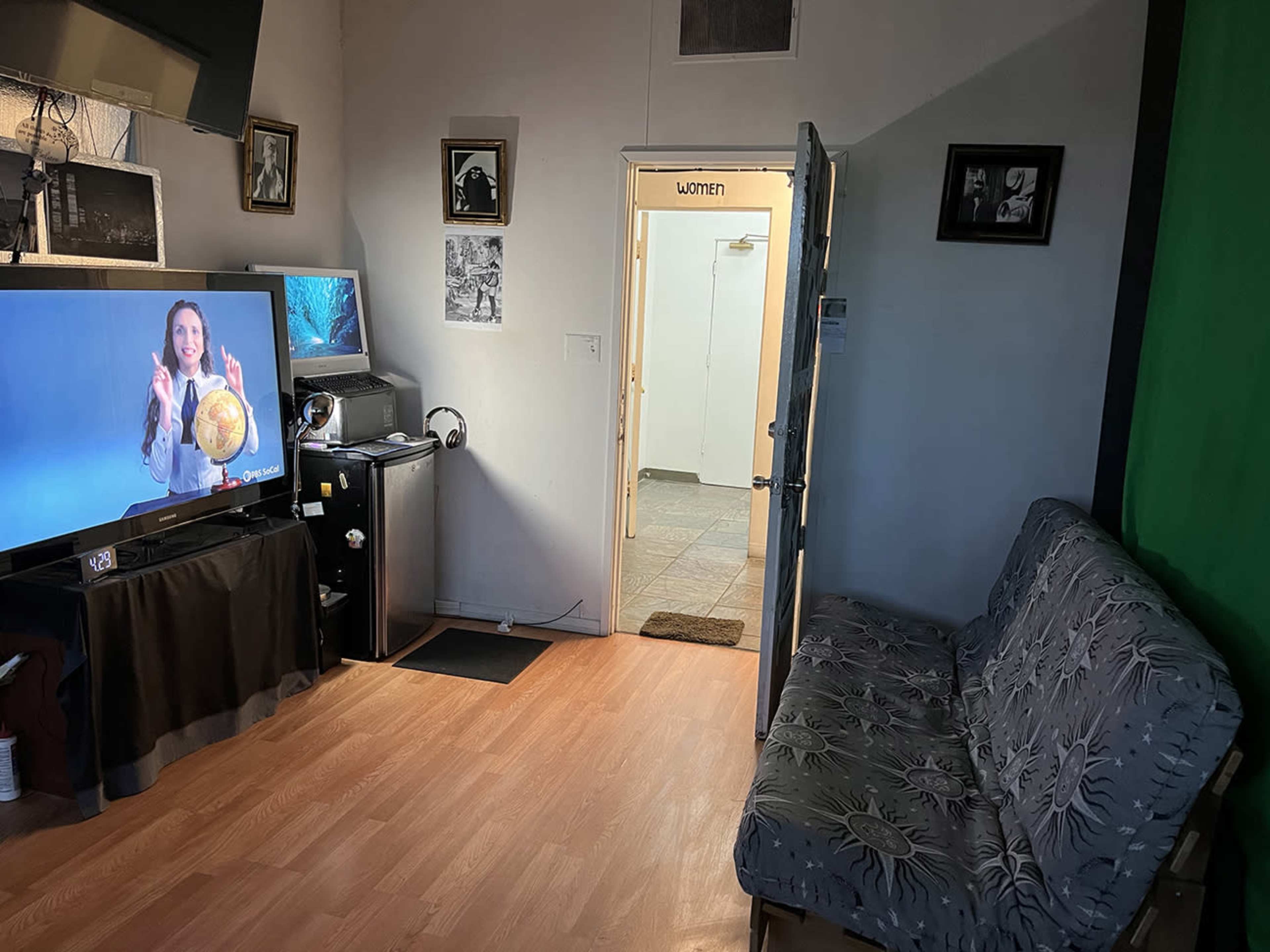 A room with a television displaying a woman gesturing beside a sofa, with a door leading to a bathroom labeled "WOMEN" in the background.
