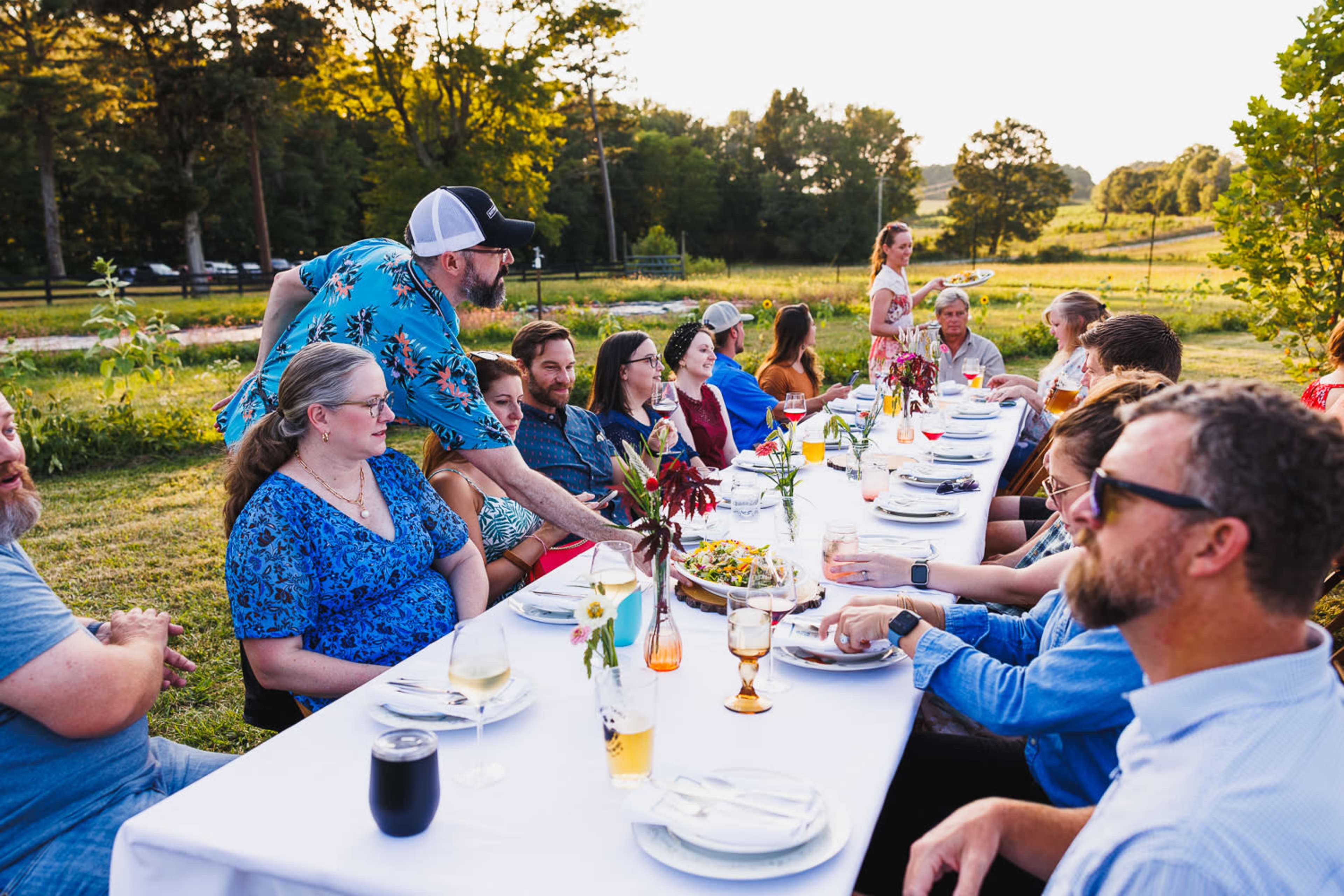 A long table is set outdoors for a gathering, with people seated on either side, enjoying food and drinks against a backdrop of trees and a sunset.