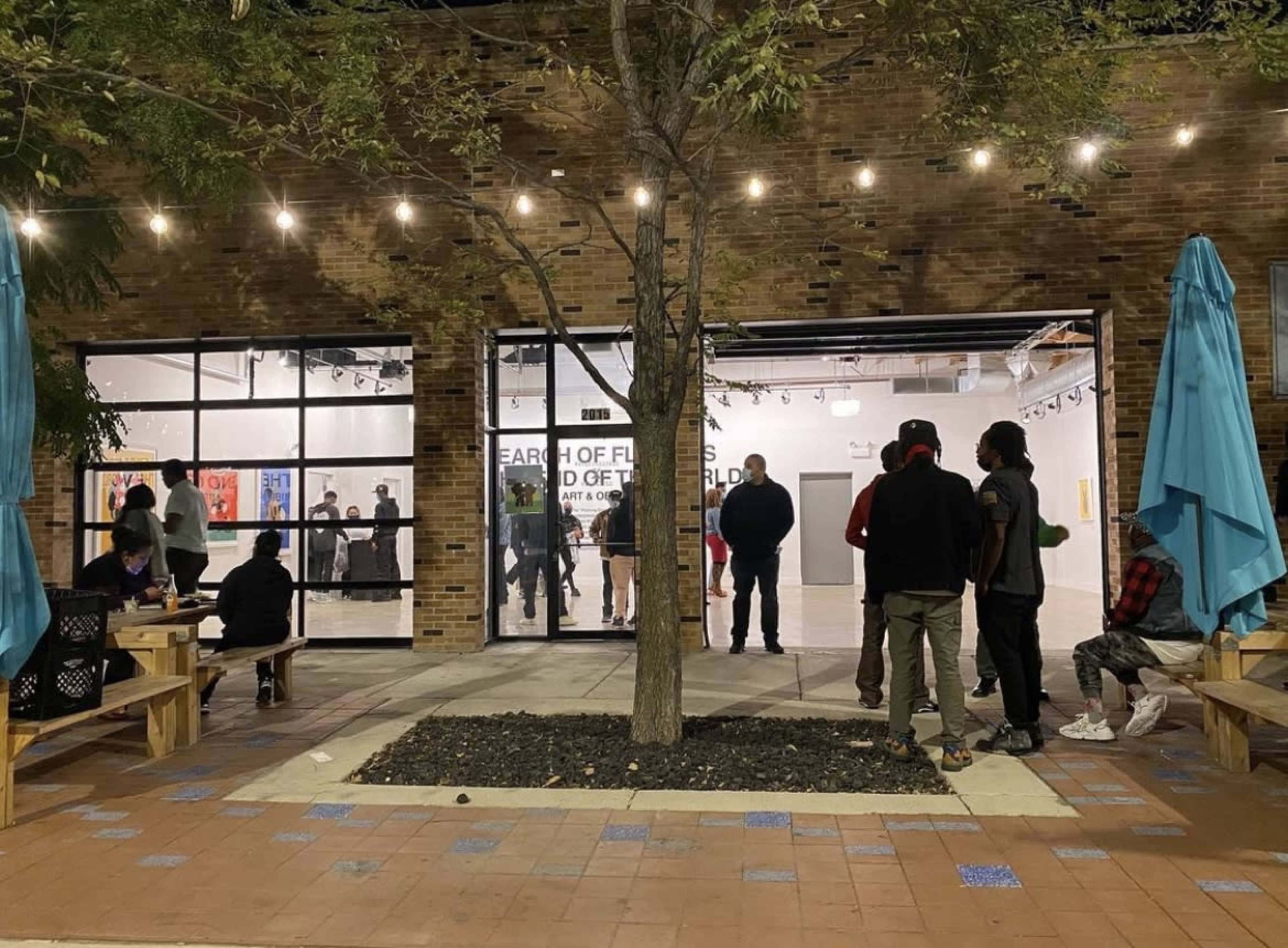 People gather outside a gallery, with some seated at benches and others standing near a tree, as warm lights illuminate the space.