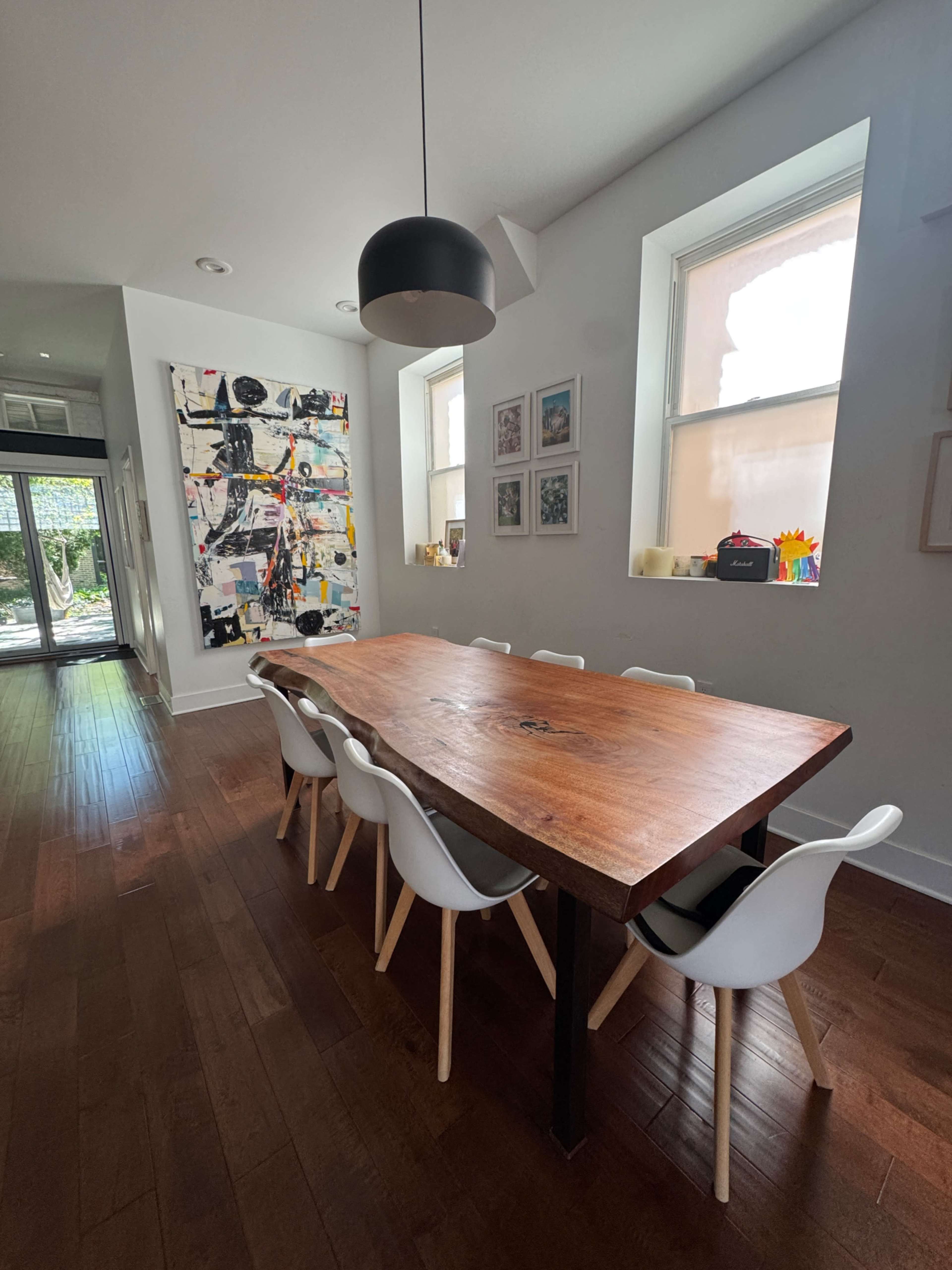 A wooden dining table with white chairs is positioned in a bright room featuring large windows and colorful artwork on the walls.