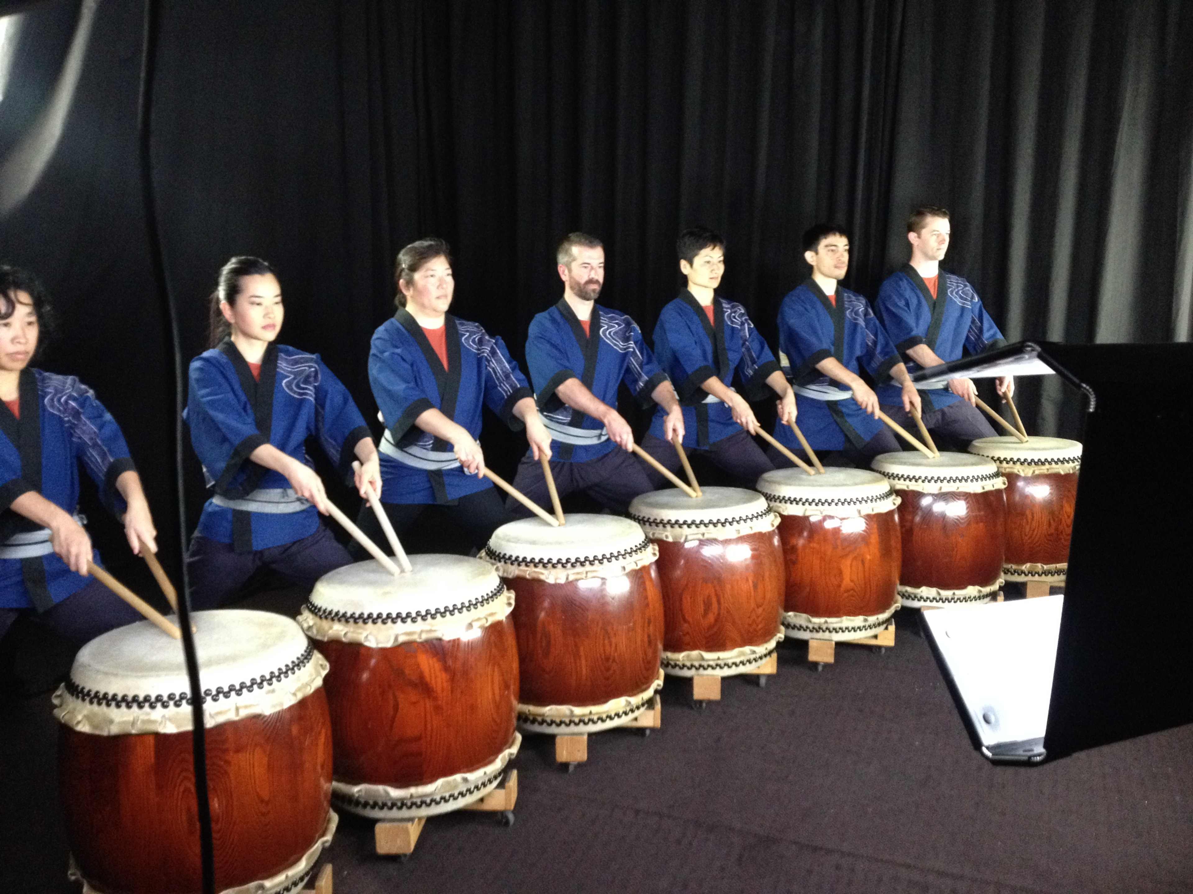 A group of seven individuals wearing blue uniforms plays large taiko drums in front of a black backdrop.