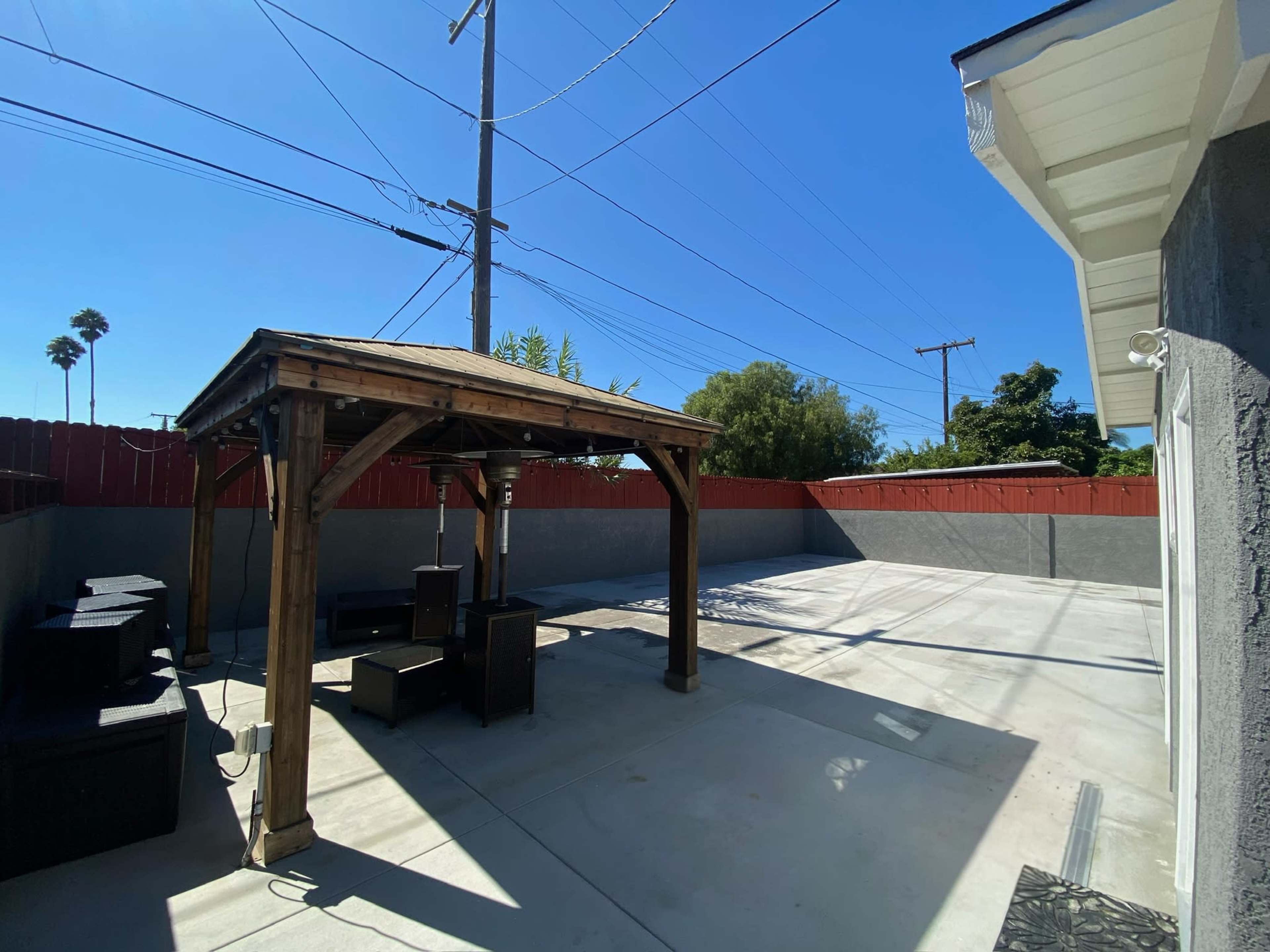 A wooden gazebo stands in an empty concrete yard, enclosed by a red fence and power lines against a clear blue sky.