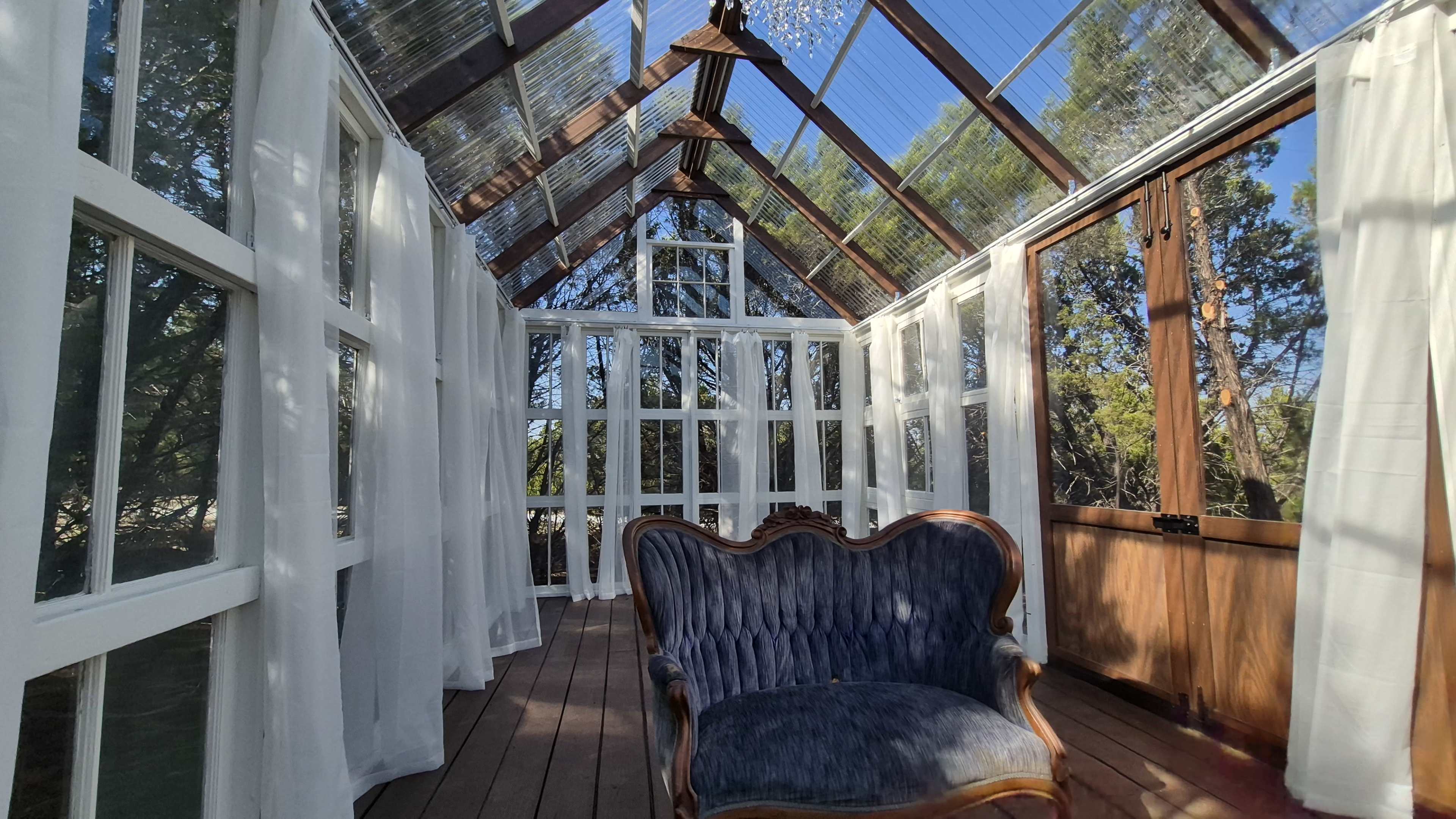 The image shows a sunlit greenhouse interior with a vintage upholstered chair and large windows allowing light to filter through.