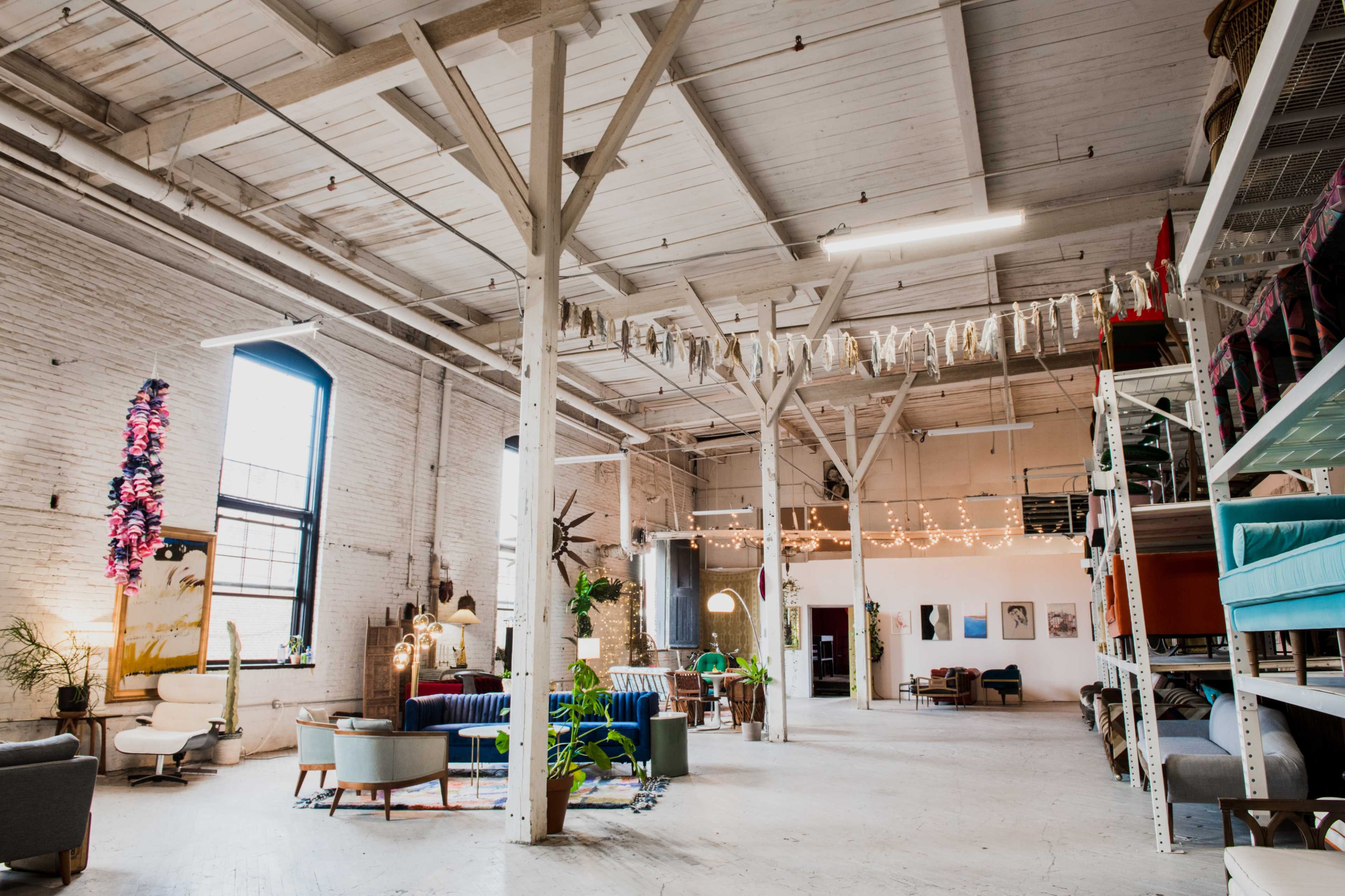 A spacious, industrial-style interior featuring white brick walls, high ceilings, wooden beams, and various seating arrangements with decorative art and plants.