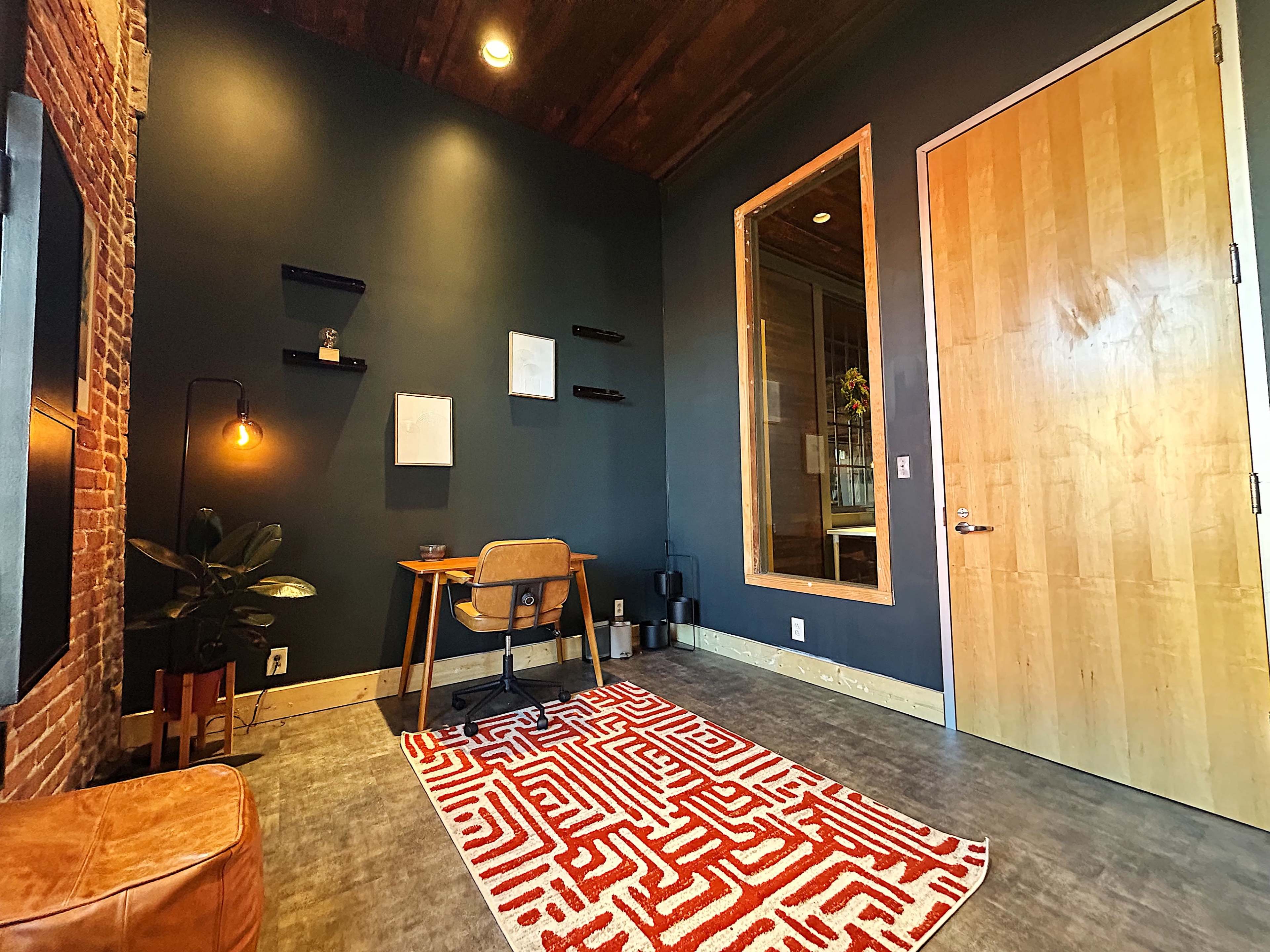The image shows a small, modern workspace featuring a desk with a chair, a large window, and a textured area rug on a concrete floor.