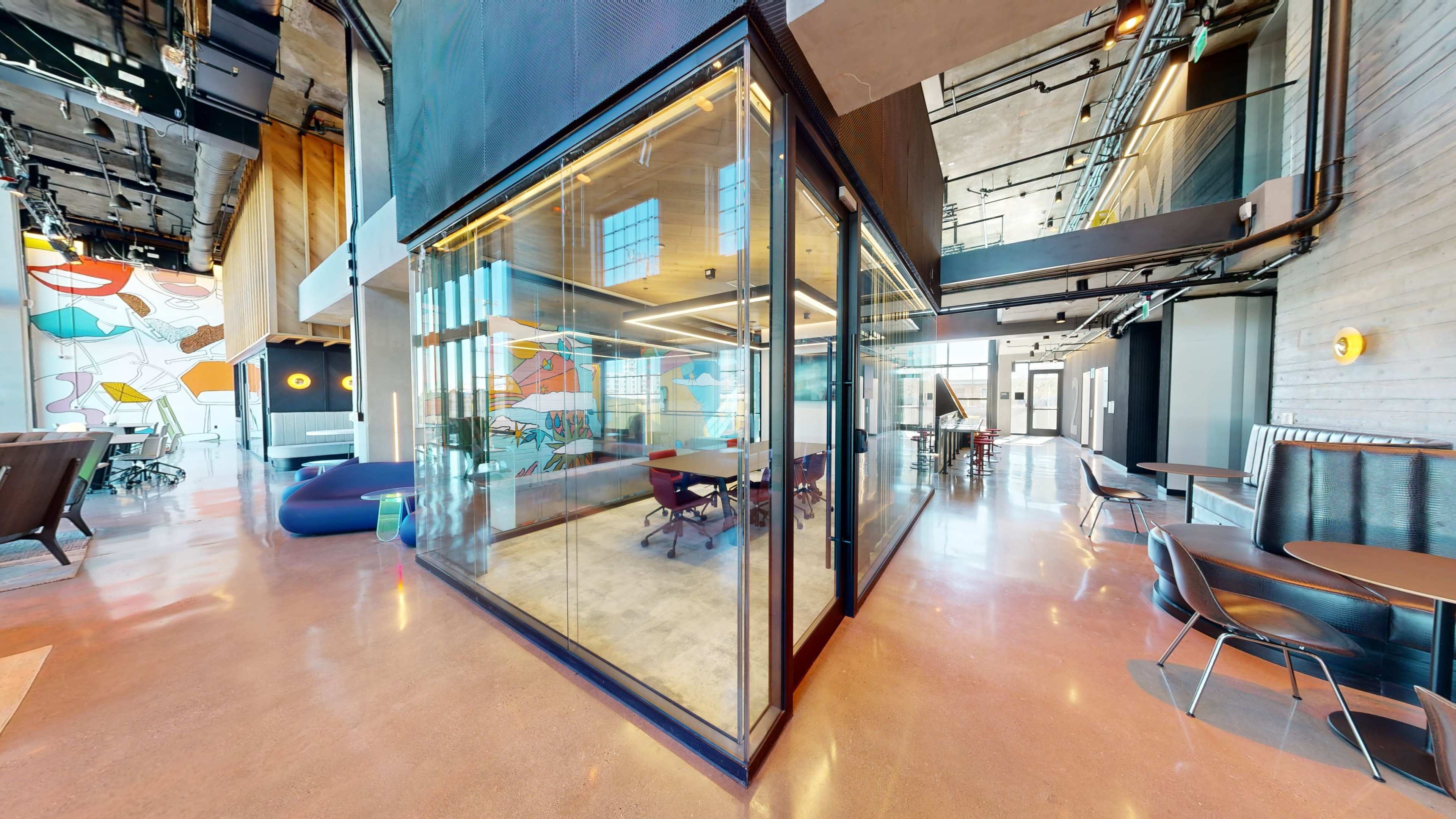 The image shows a modern office space featuring a glass-walled conference room, open seating areas, and colorful wall art.