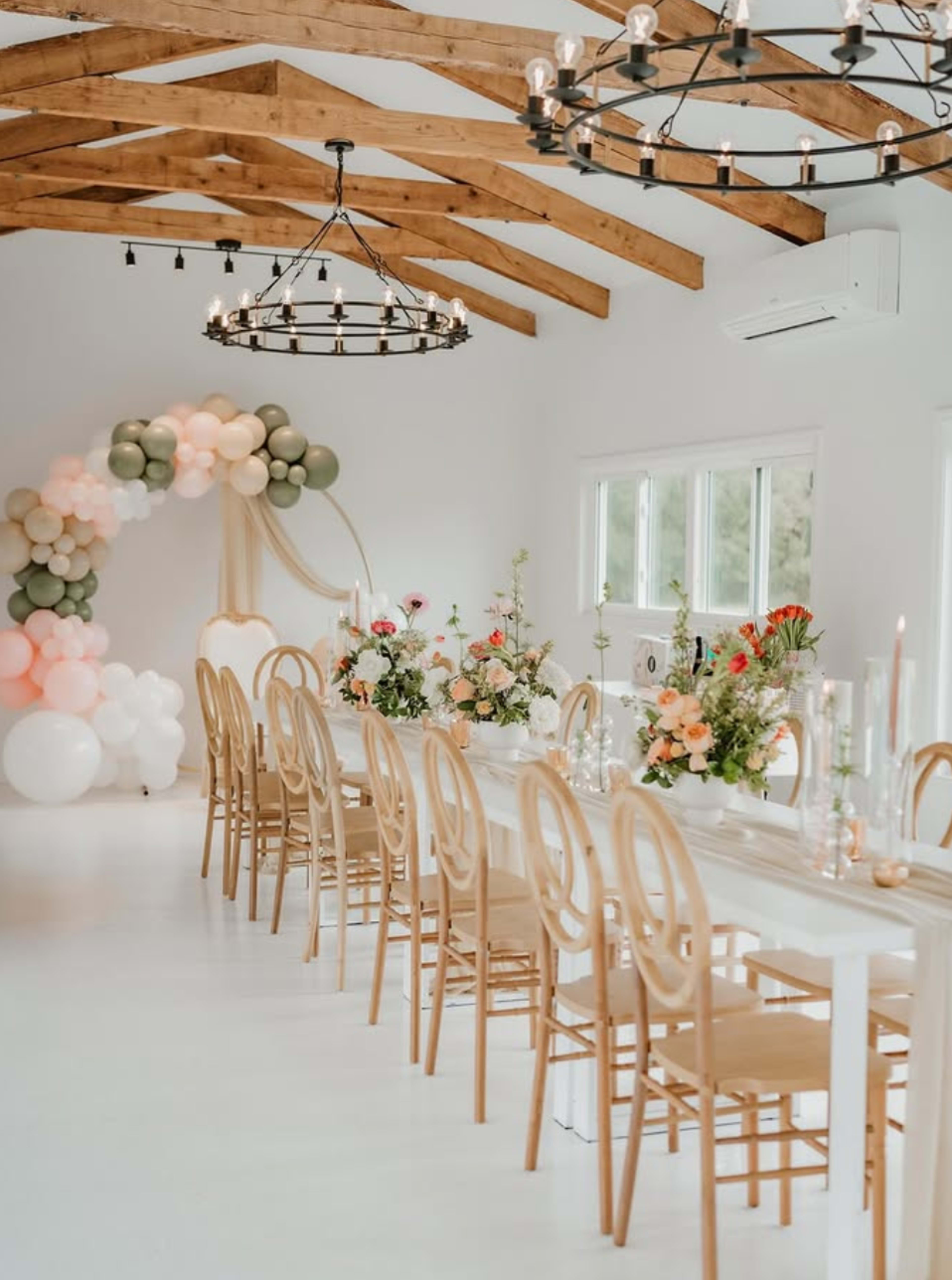 A long banquet table decorated with flowers and candles is set in a bright room with wooden beams and a balloon arch in the background.