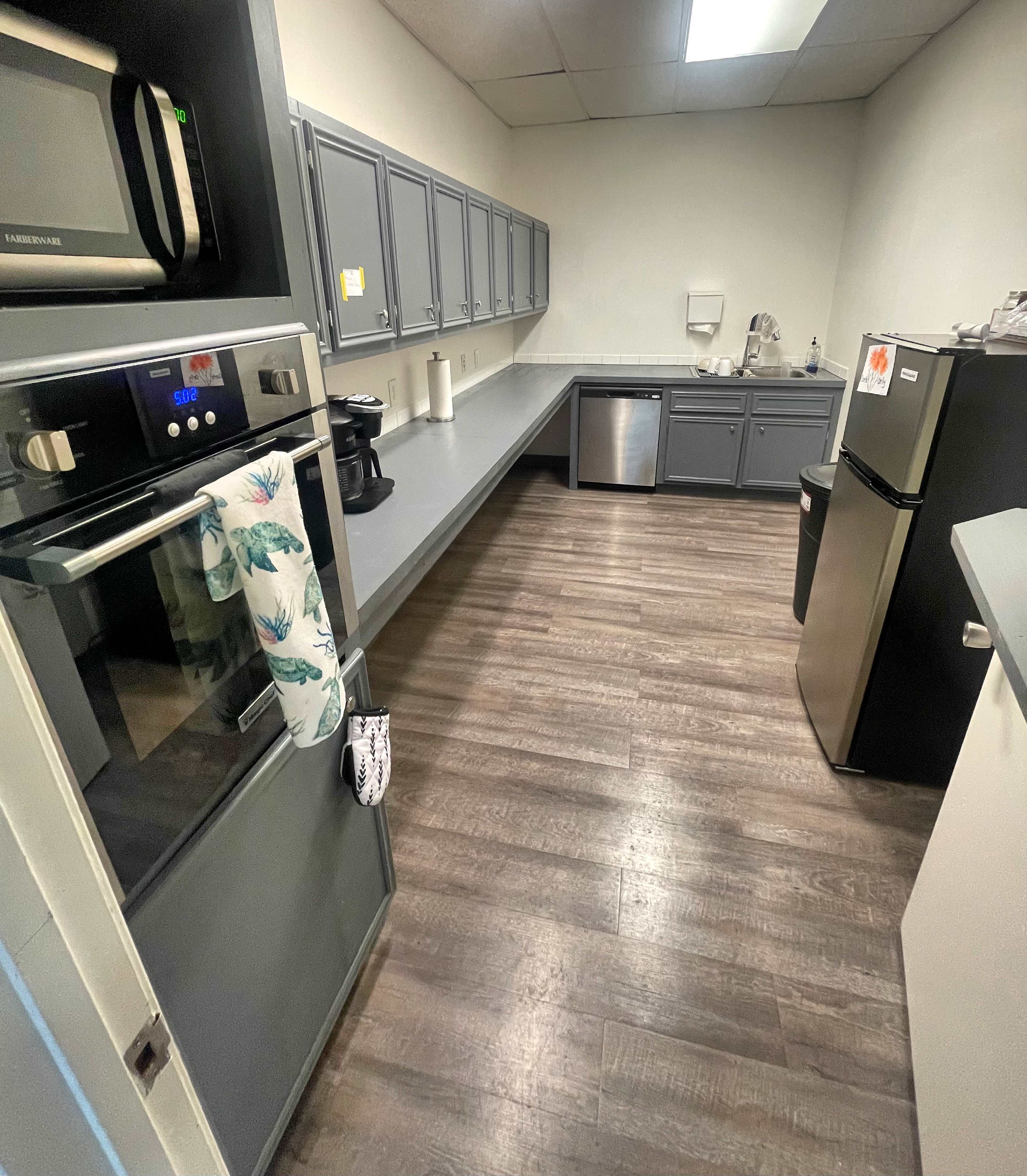 The image shows a clean kitchen with gray cabinets, a stainless steel refrigerator, and a microwave, featuring a spacious counter area and wood-like flooring.