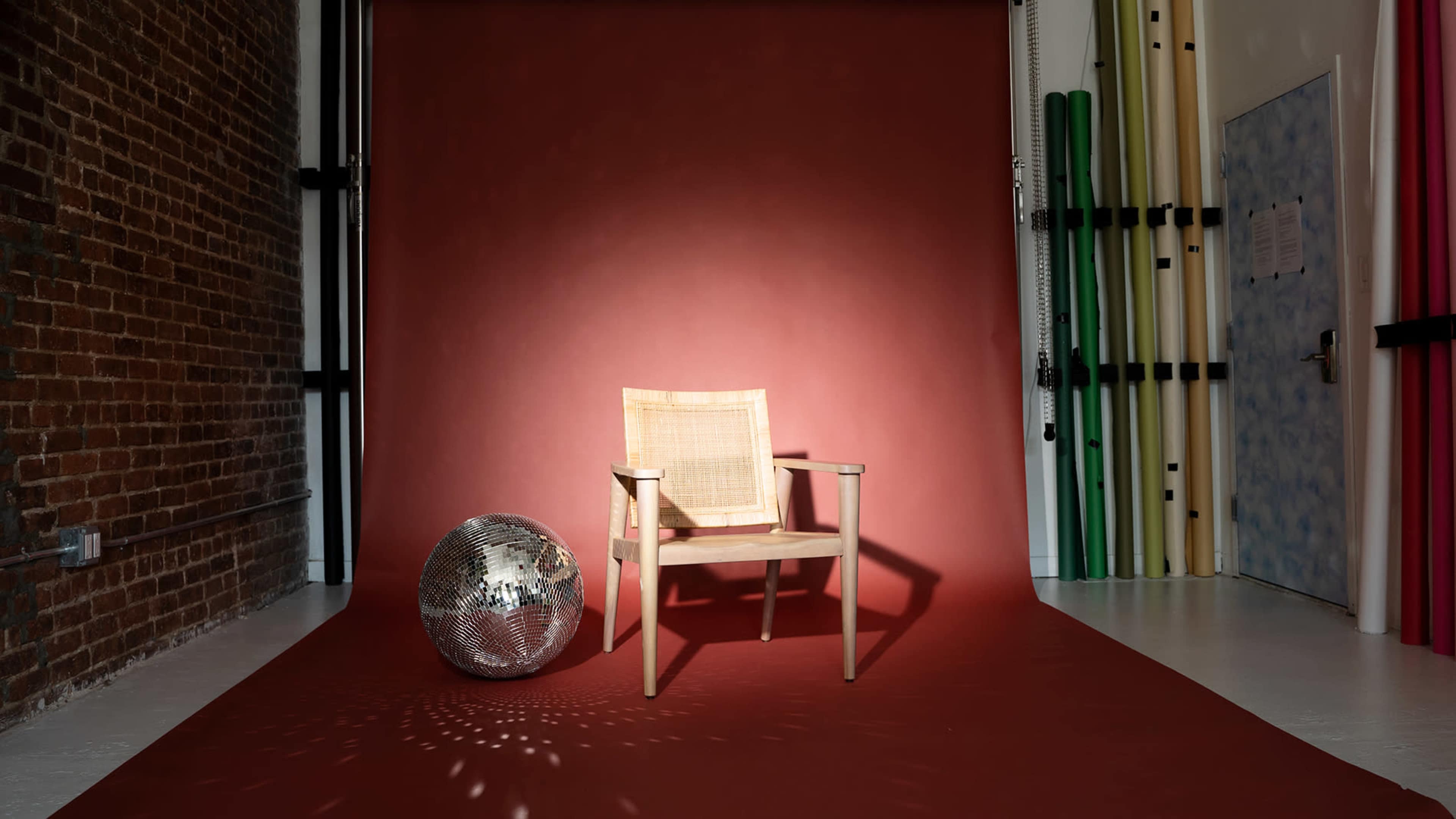 A wooden chair sits in front of a maroon backdrop beside a reflective disco ball on a studio floor.