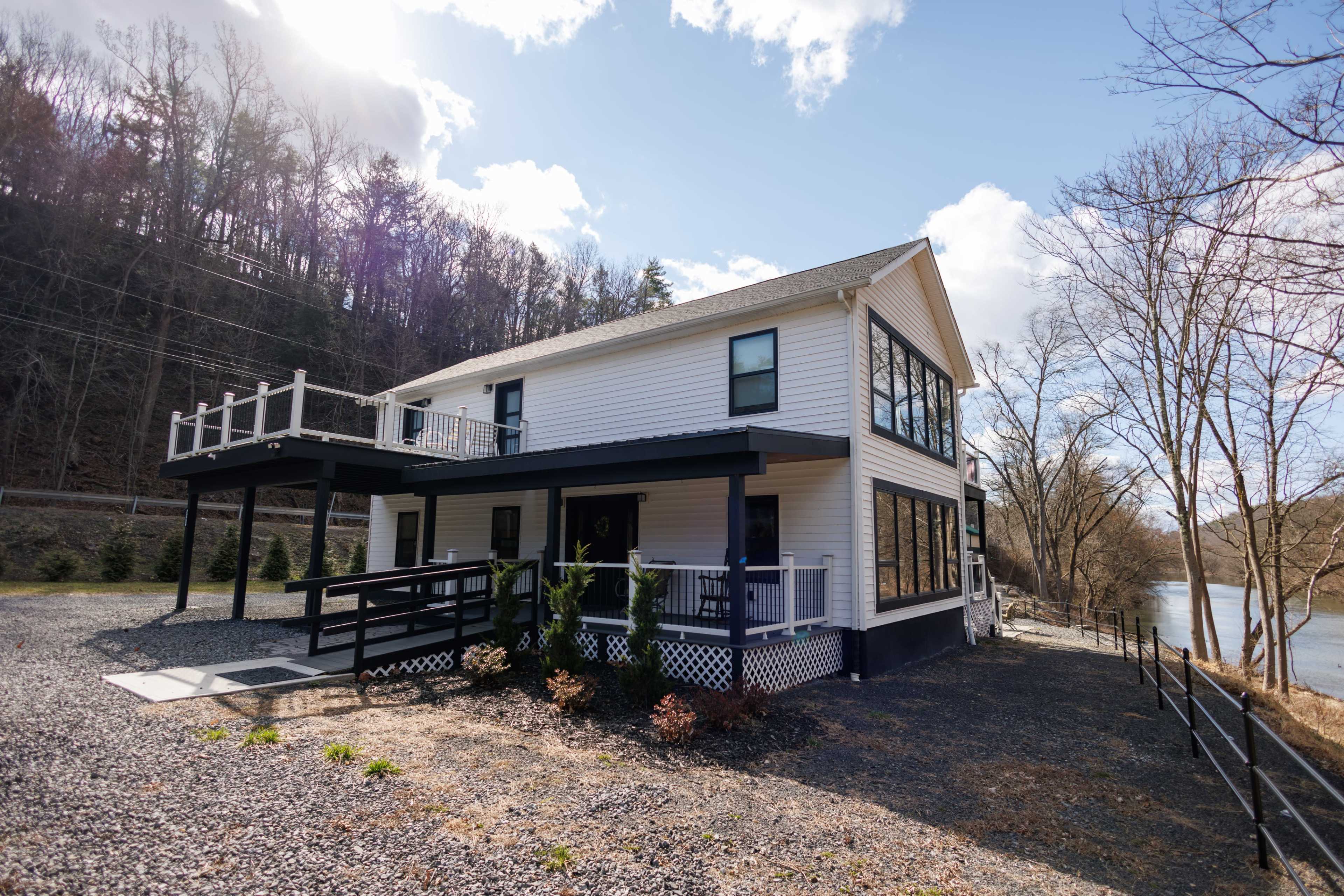 A modern two-story white house with large windows and a balcony, situated near a gravel driveway alongside a river.