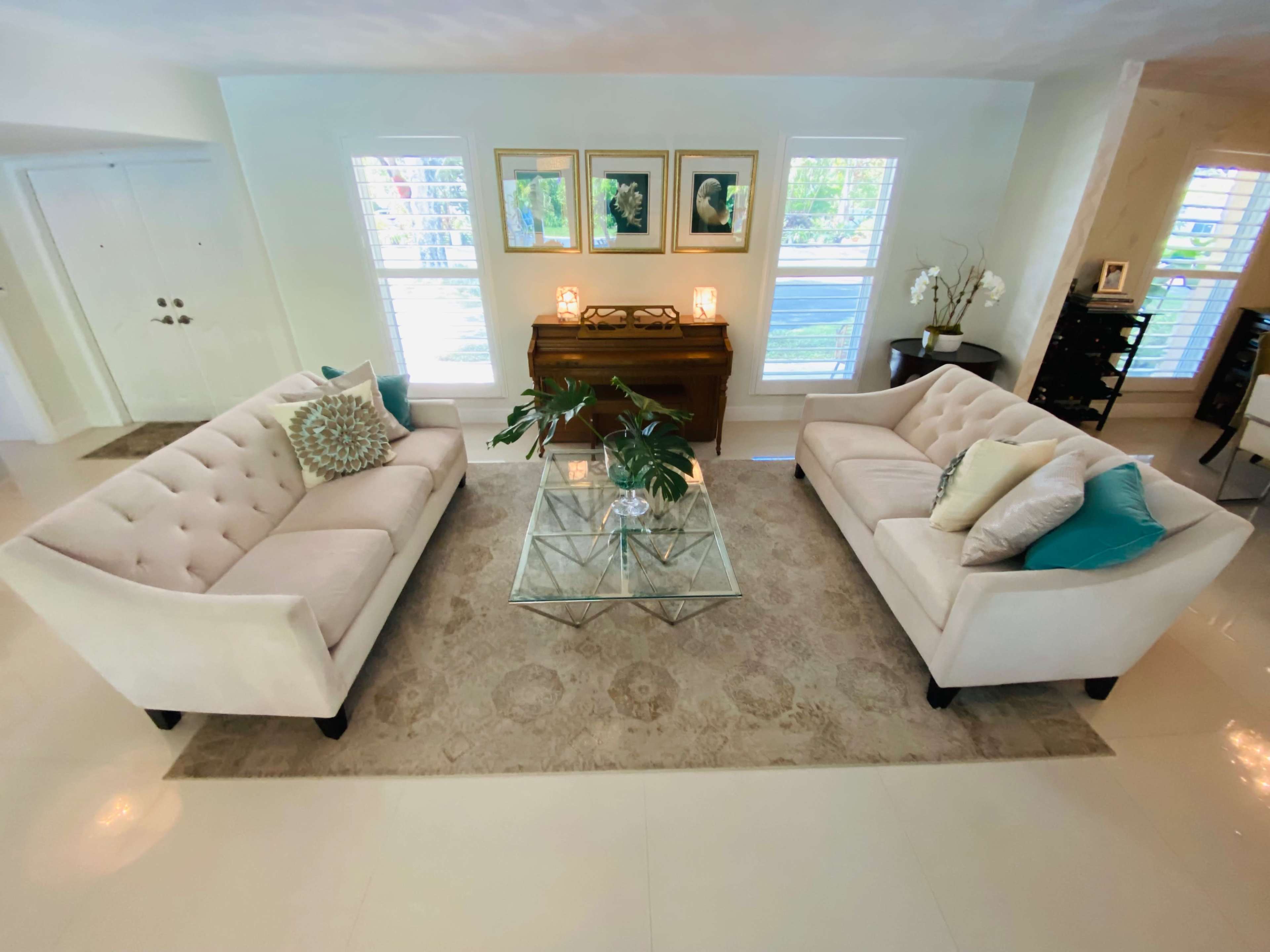 The image shows a bright living room with two tufted sofas facing each other, a glass coffee table in the center, and a piano against the wall.