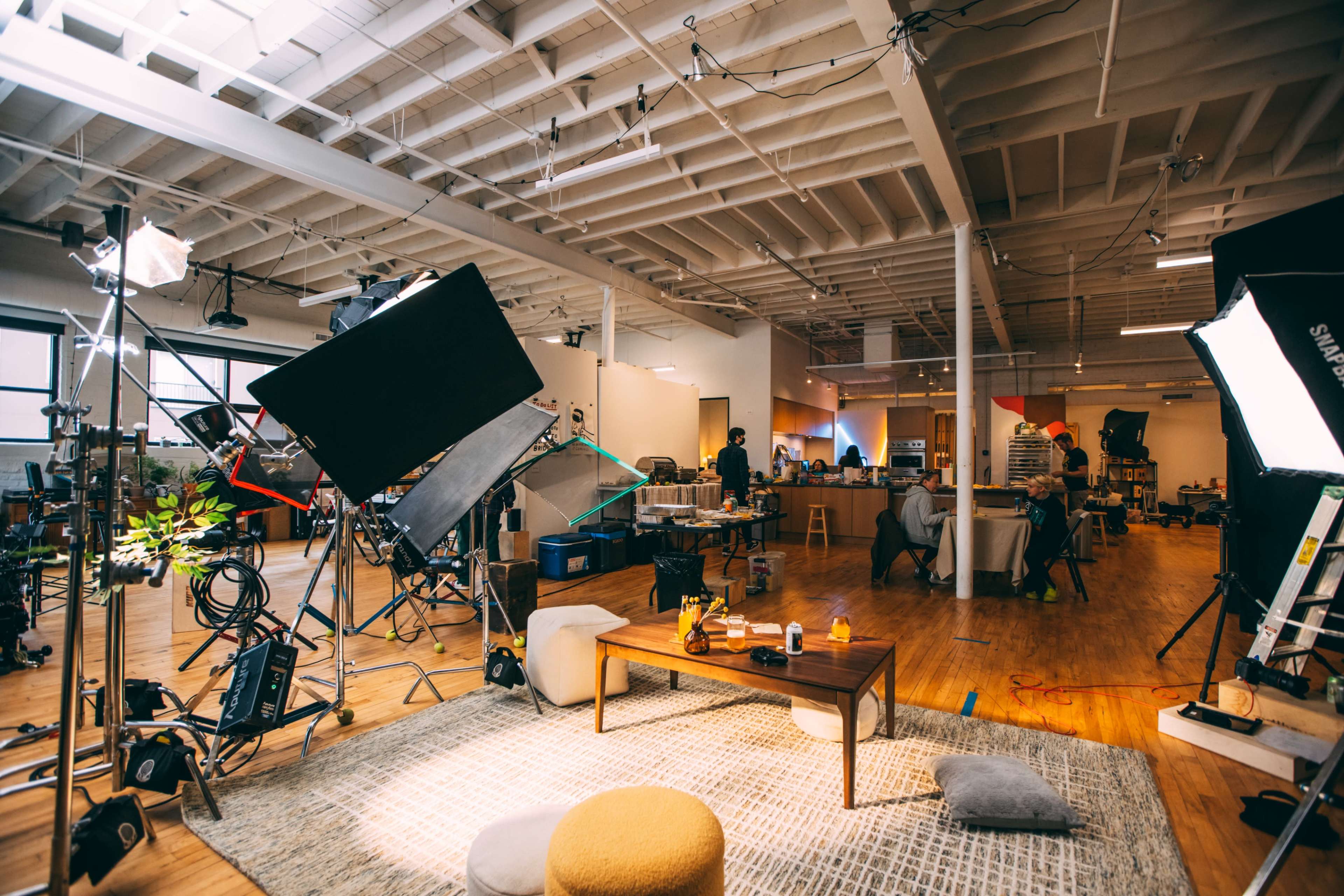 The image shows a spacious studio with photography lighting equipment, a coffee table, and several people seated in the background.