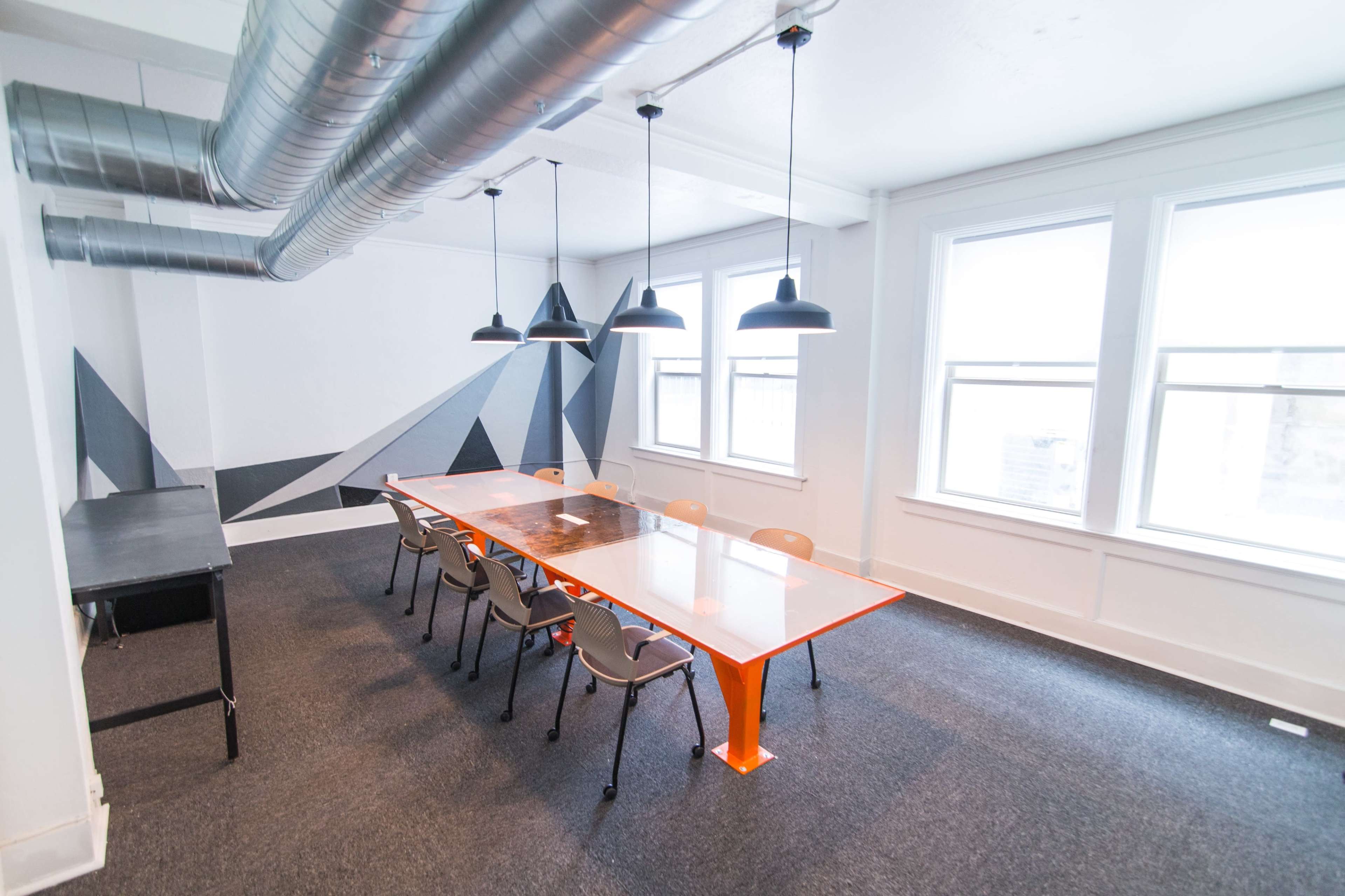 A spacious conference room features a long glass table with orange legs, surrounded by eight gray chairs, and is illuminated by pendant lights with a geometric mural on the wall.