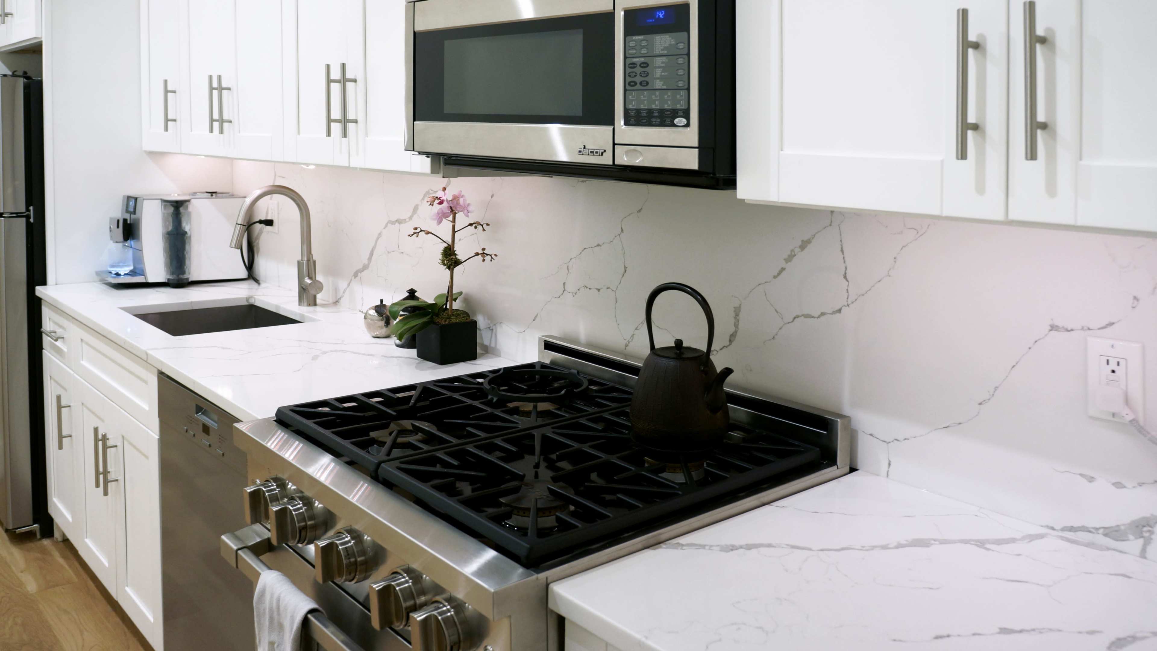 A modern kitchen features a stovetop with a black kettle, white cabinetry, a microwave above, and a marble countertop with a small potted orchid.