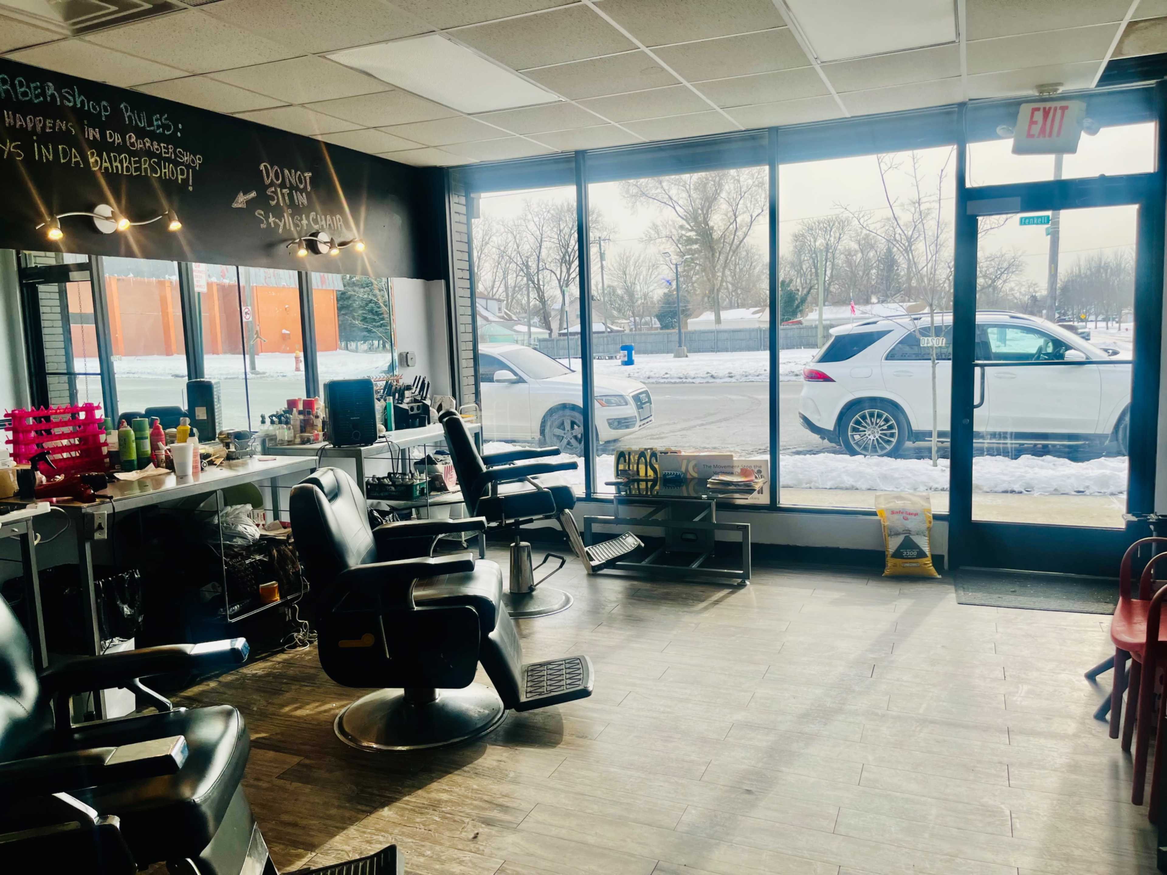 The interior of a barbershop features two black barber chairs, mirrors, and a large window overlooking a snowy street.