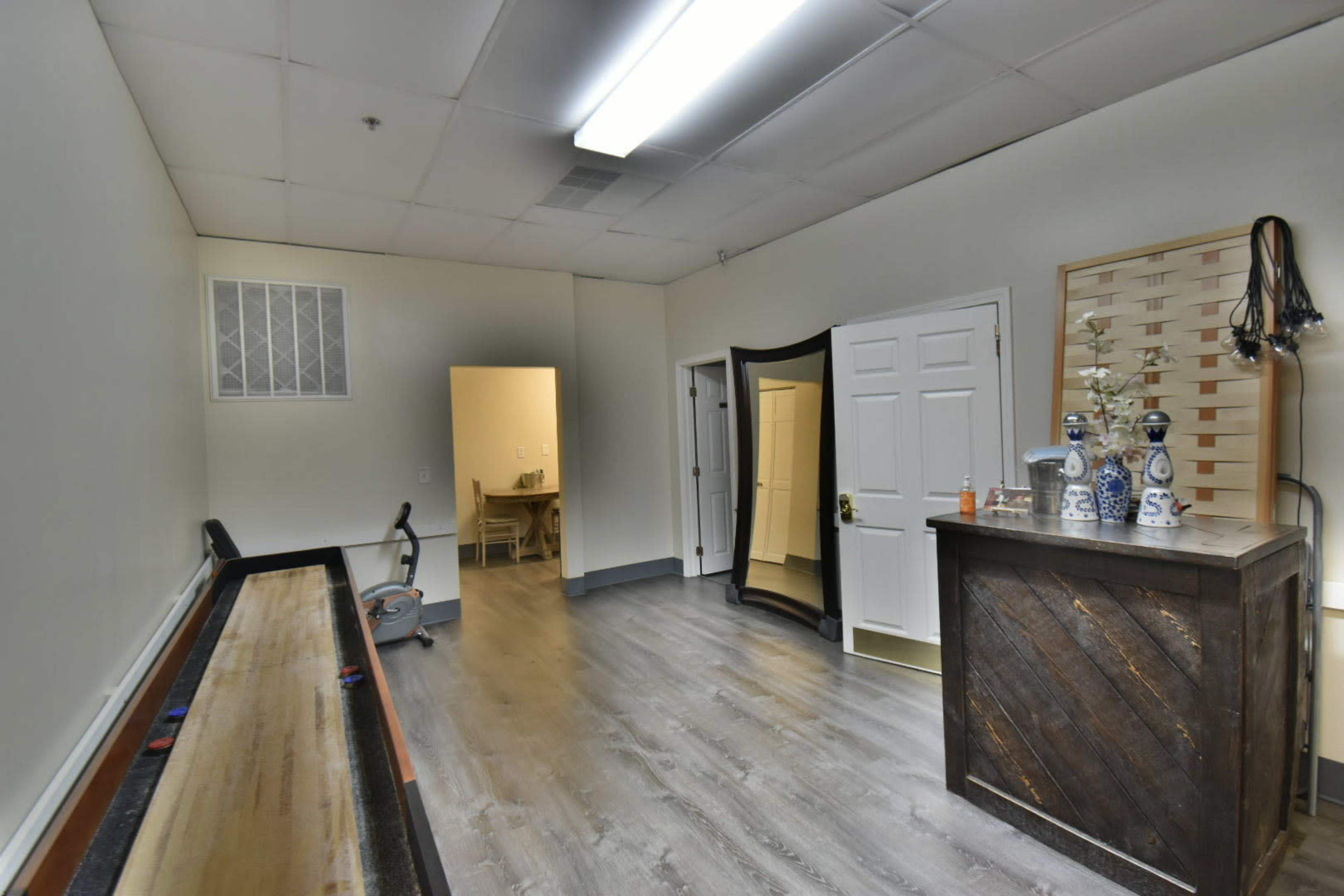 The image shows a spacious, well-lit room with a wooden counter, a large mirror, and a long wooden table along one wall, leading to a doorway at the back.