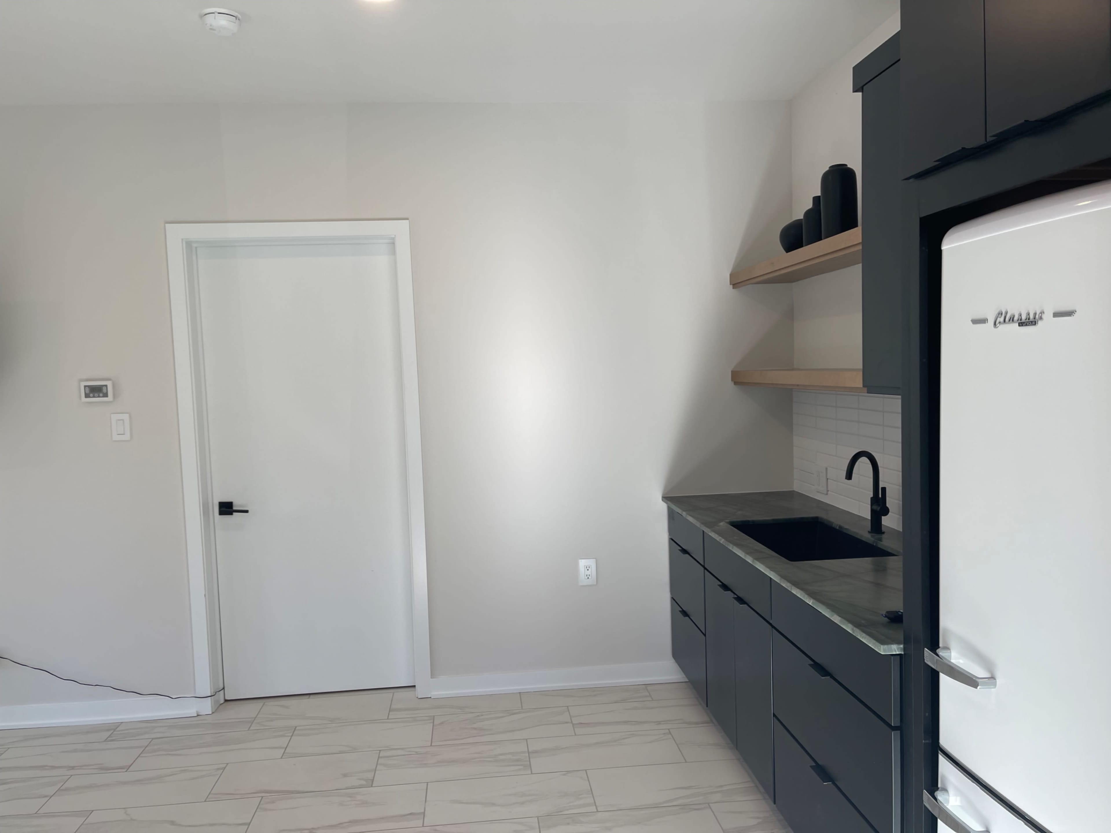 The image shows a modern kitchenette with a sink, dark cabinetry, open shelves, and a white refrigerator against a plain wall with a door.