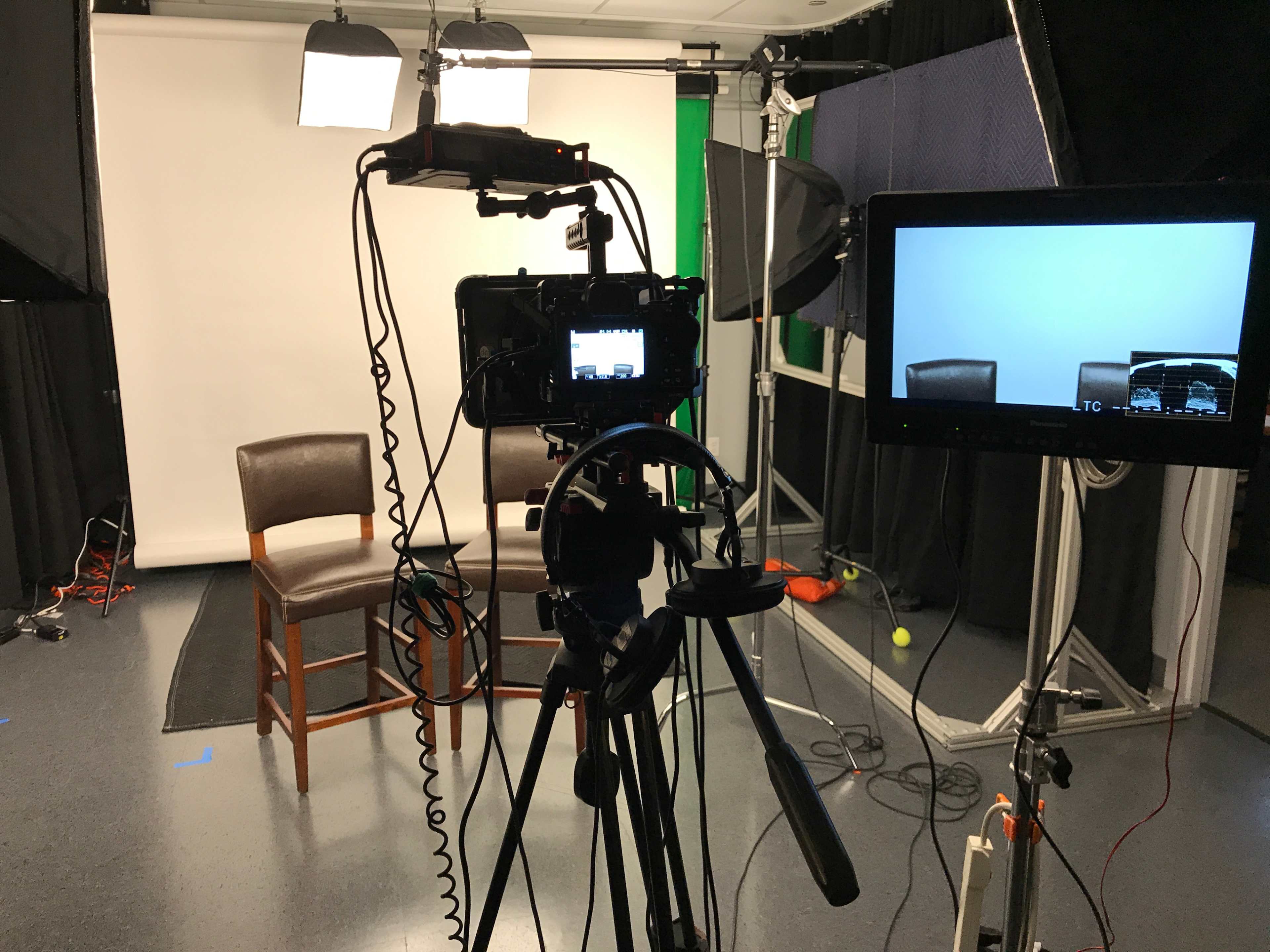 A video recording setup with a camera on a tripod facing two empty chairs in a studio environment, surrounded by lighting equipment and a green screen.