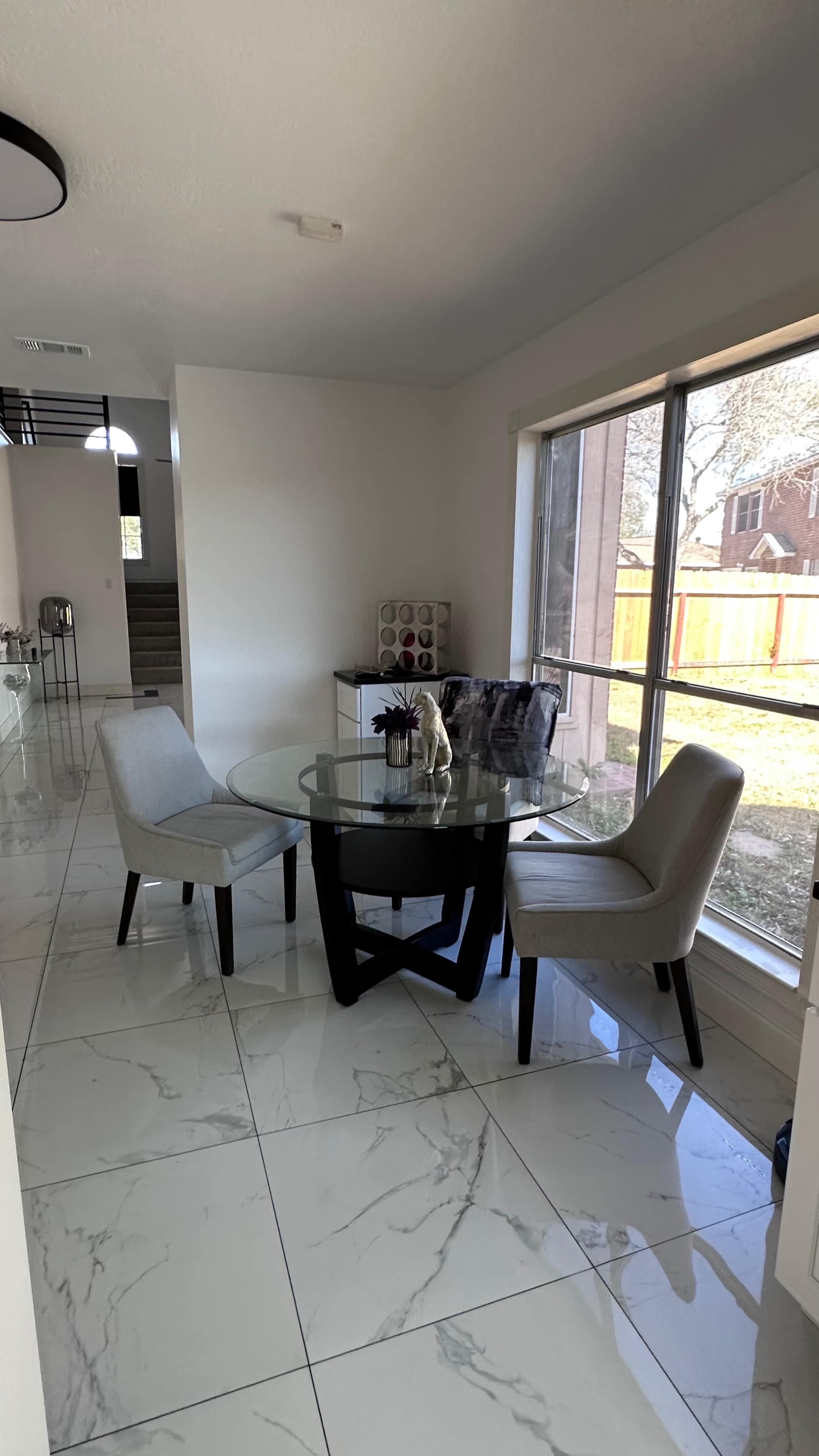 A dining area with a glass table and two upholstered chairs beside a large window overlooking an outdoor space.