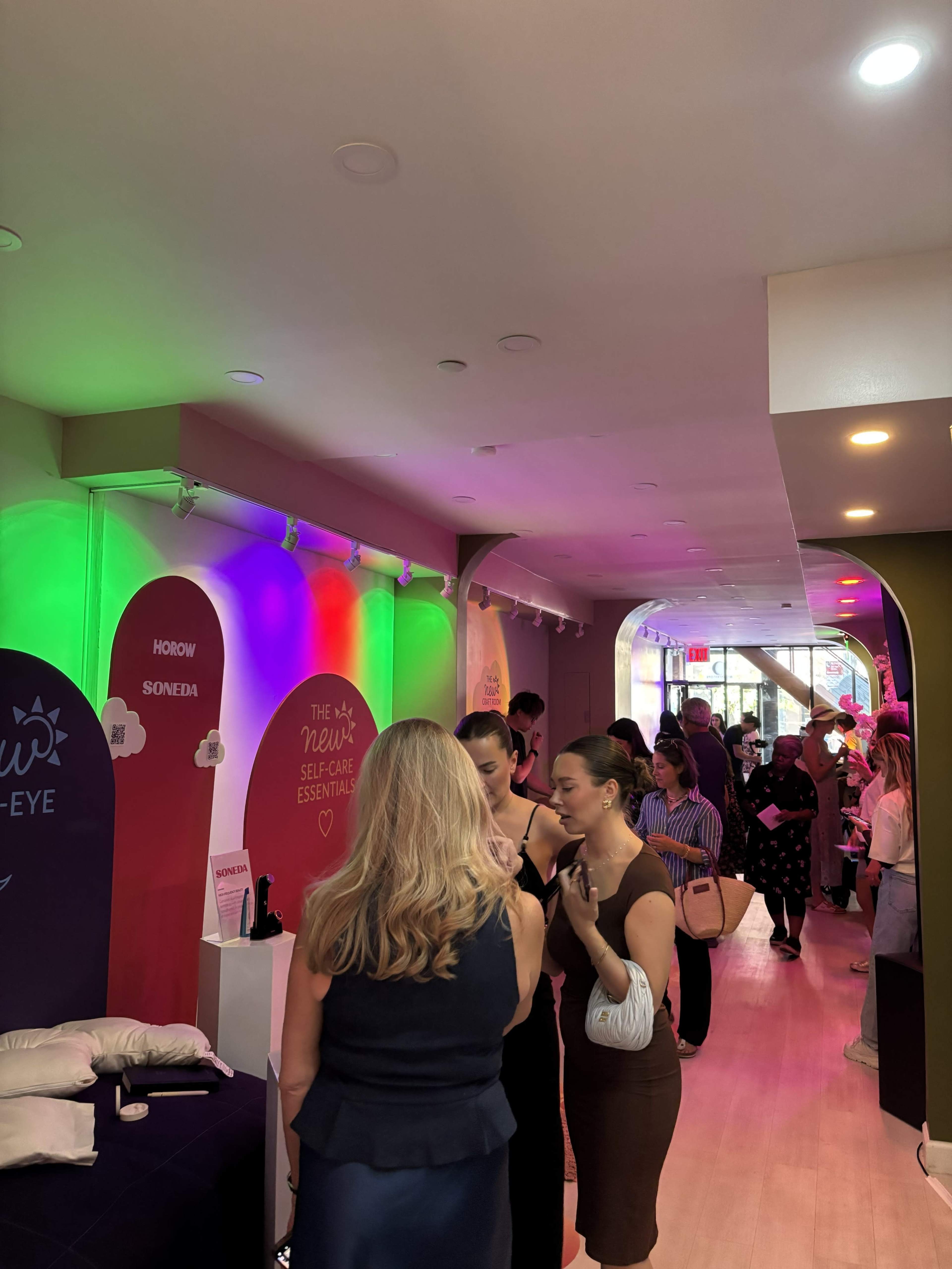 The image shows a brightly lit indoor space with colorful lighting, where a group of people are engaged in conversation, while a line of attendees waits nearby.