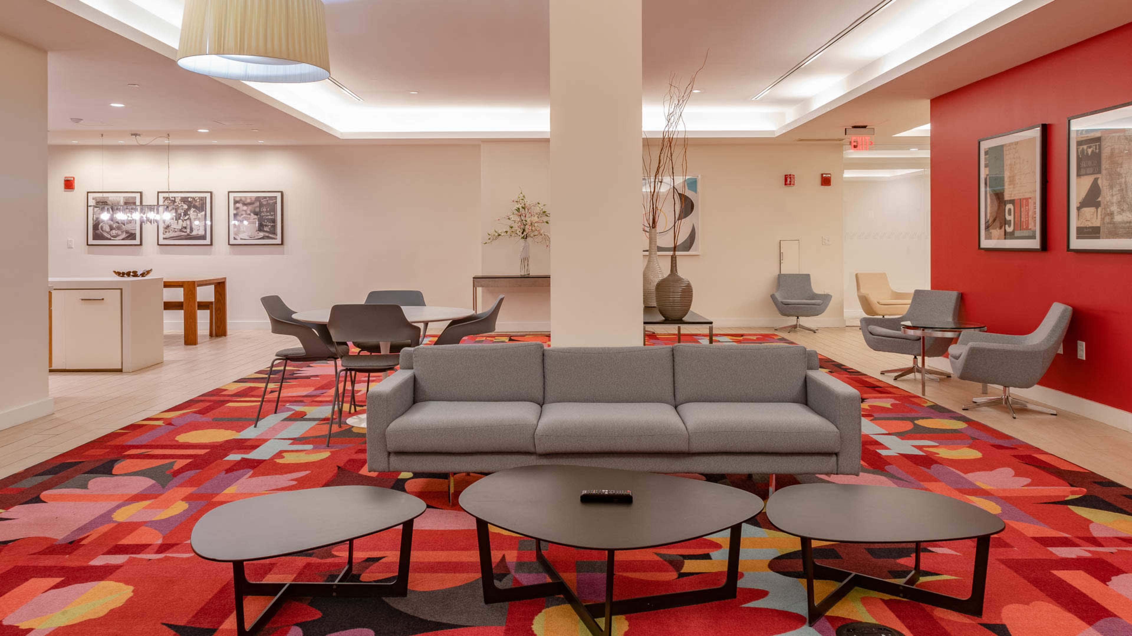 The image shows a modern lounge area with a gray couch, several small black tables, and a colorful rug, surrounded by seating and decorative wall art.