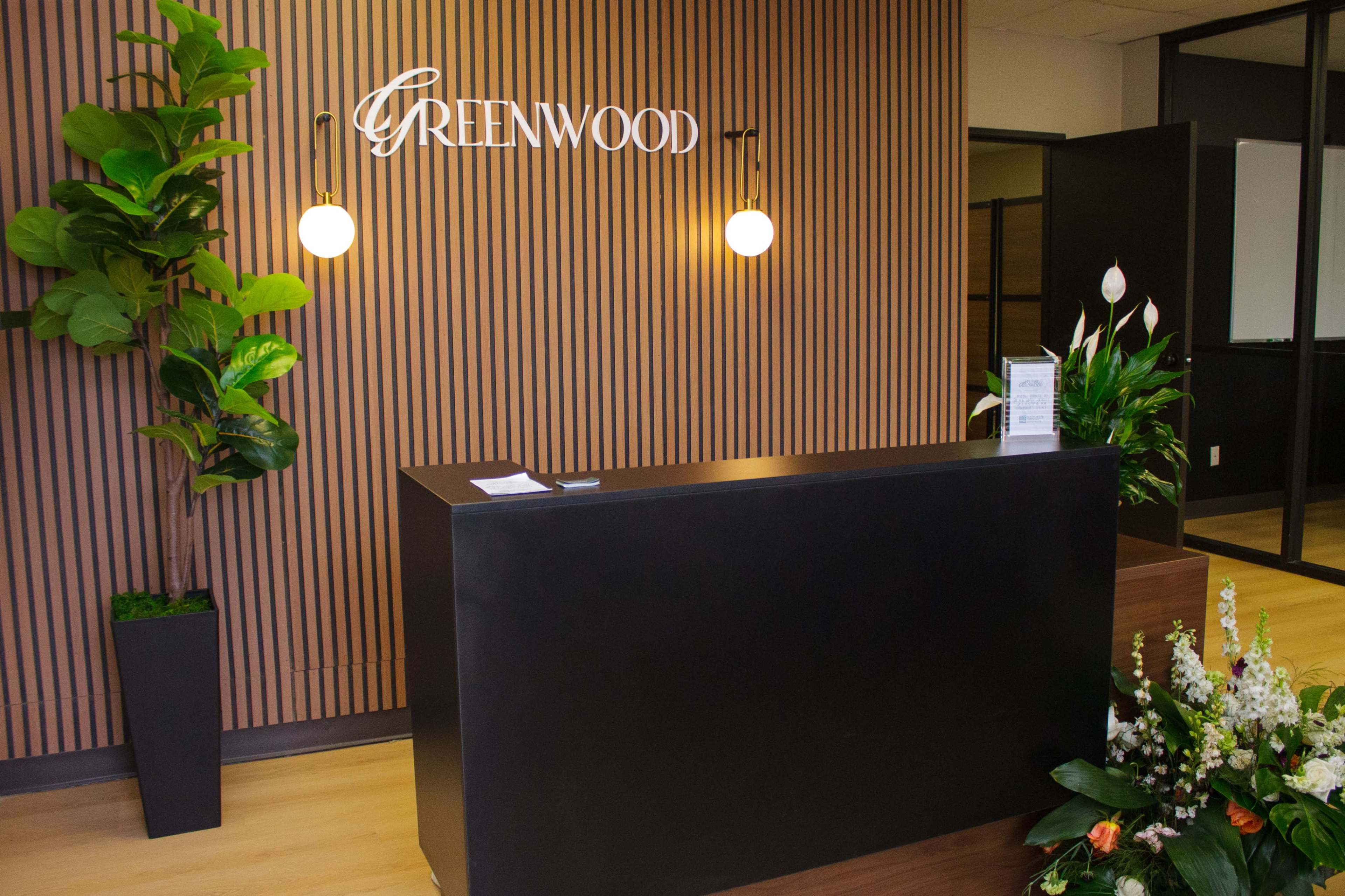 The image shows a modern reception area with a dark counter, a wooden wall feature, and plants on either side, including a floral arrangement.