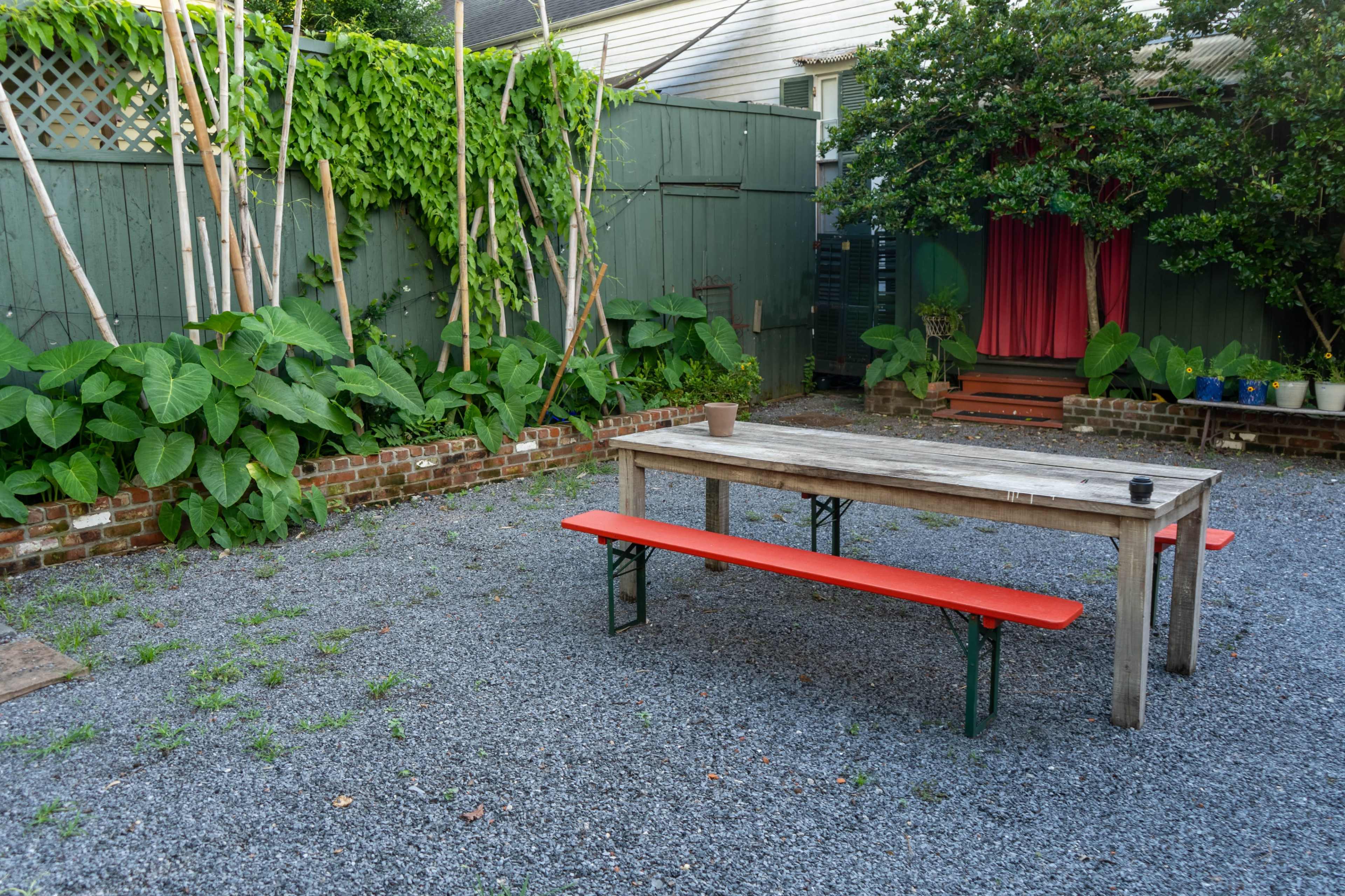 The image shows a gravel courtyard featuring a wooden table with orange benches, surrounded by green plants and a decorative backdrop.