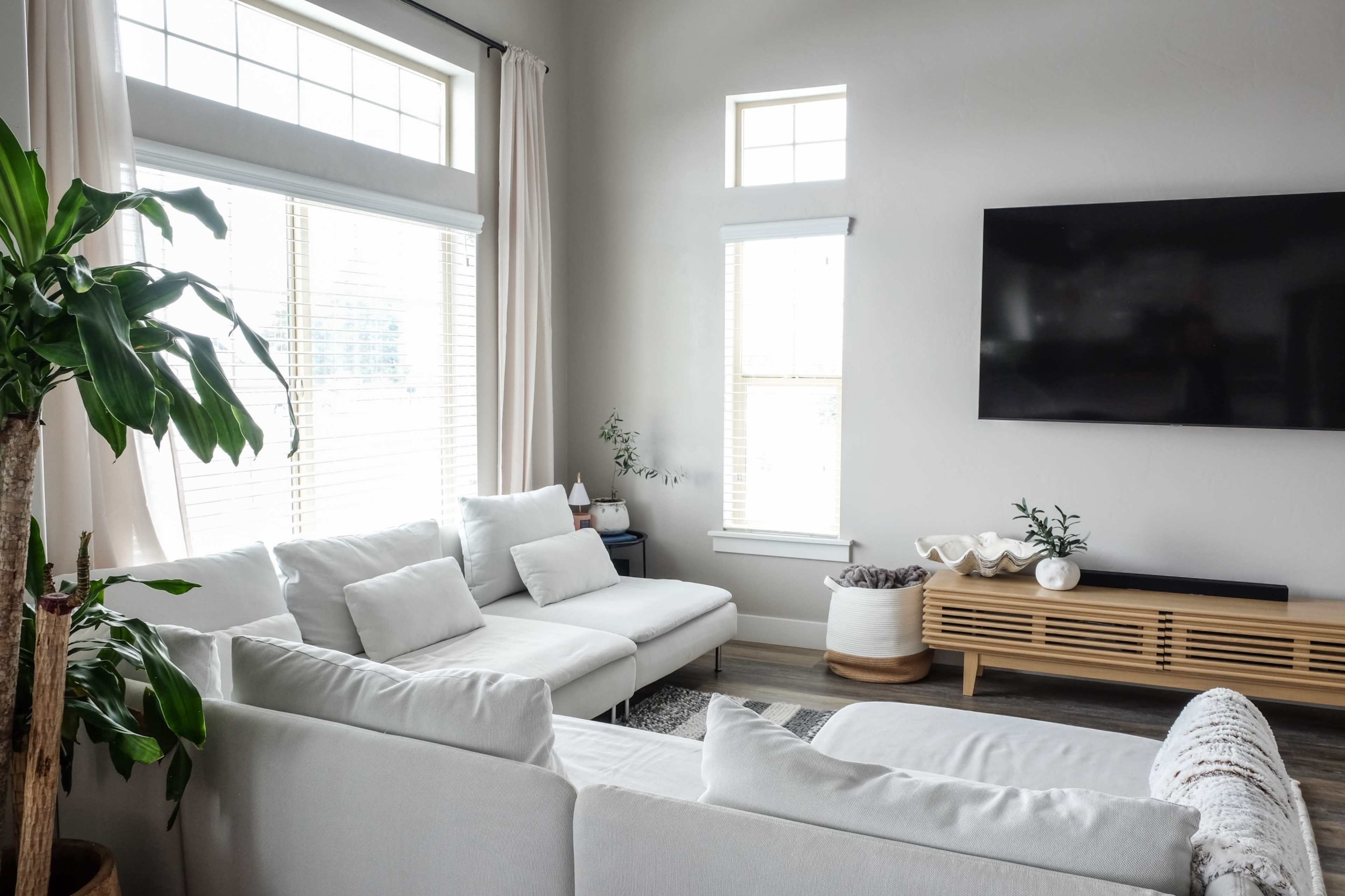 A modern living room features a white sectional sofa, a minimalist wooden media console, large windows with soft curtains, and light-colored walls.