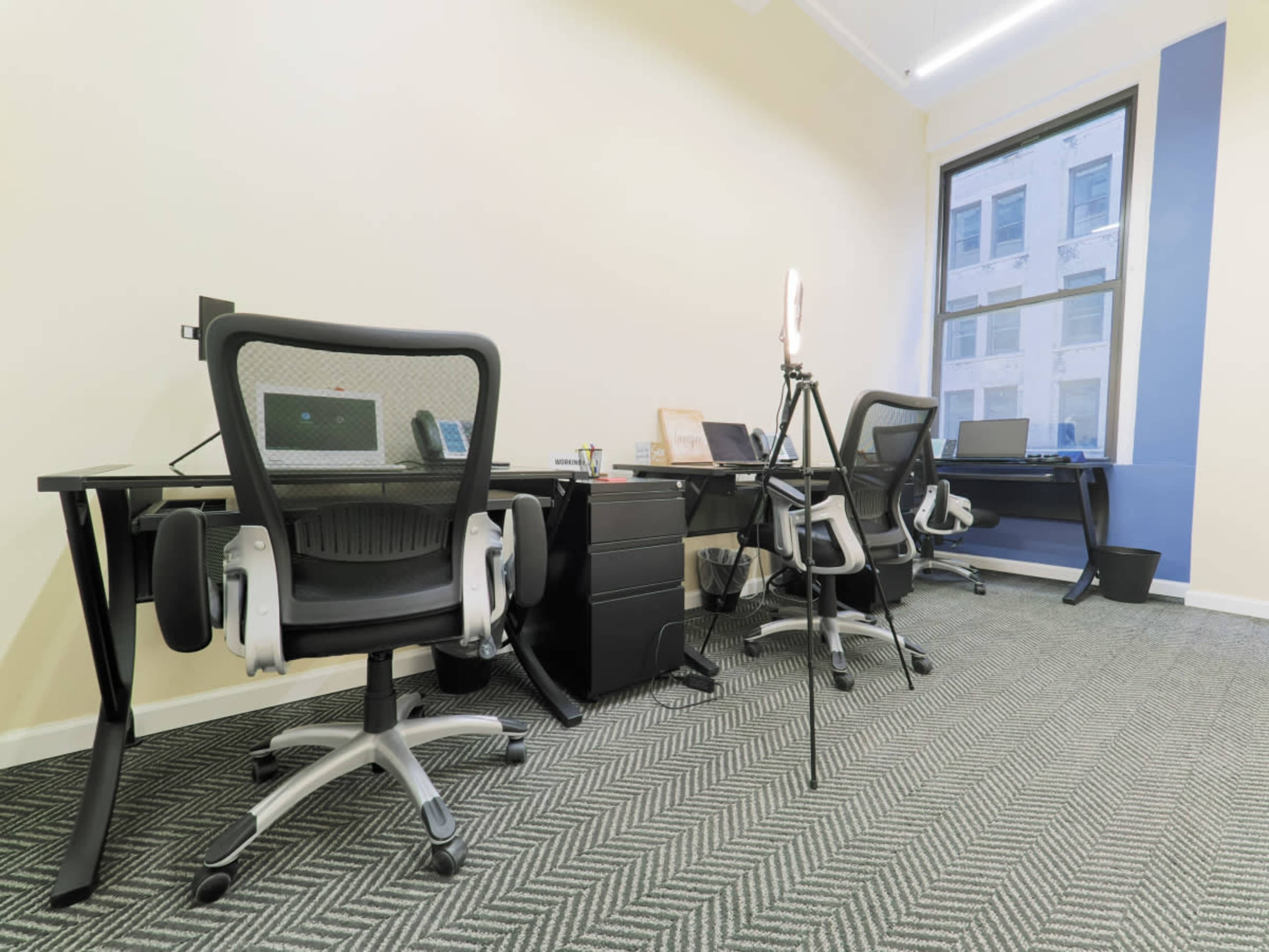 A modern office space with two black desks, each equipped with office chairs, computers, and a tripod with a ring light.