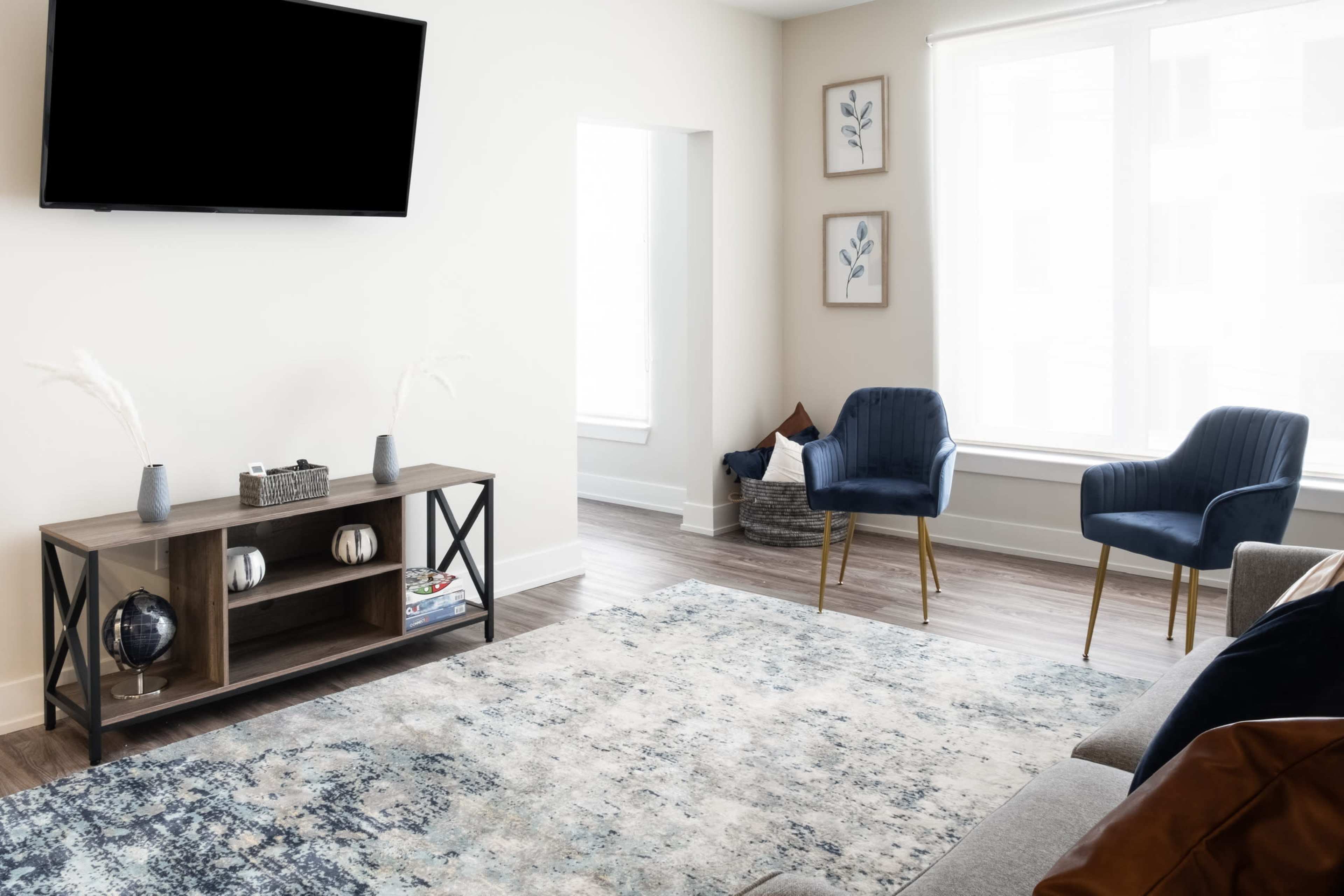 A modern living room features a television mounted on the wall, a wooden entertainment unit, two blue chairs, and a large area rug.