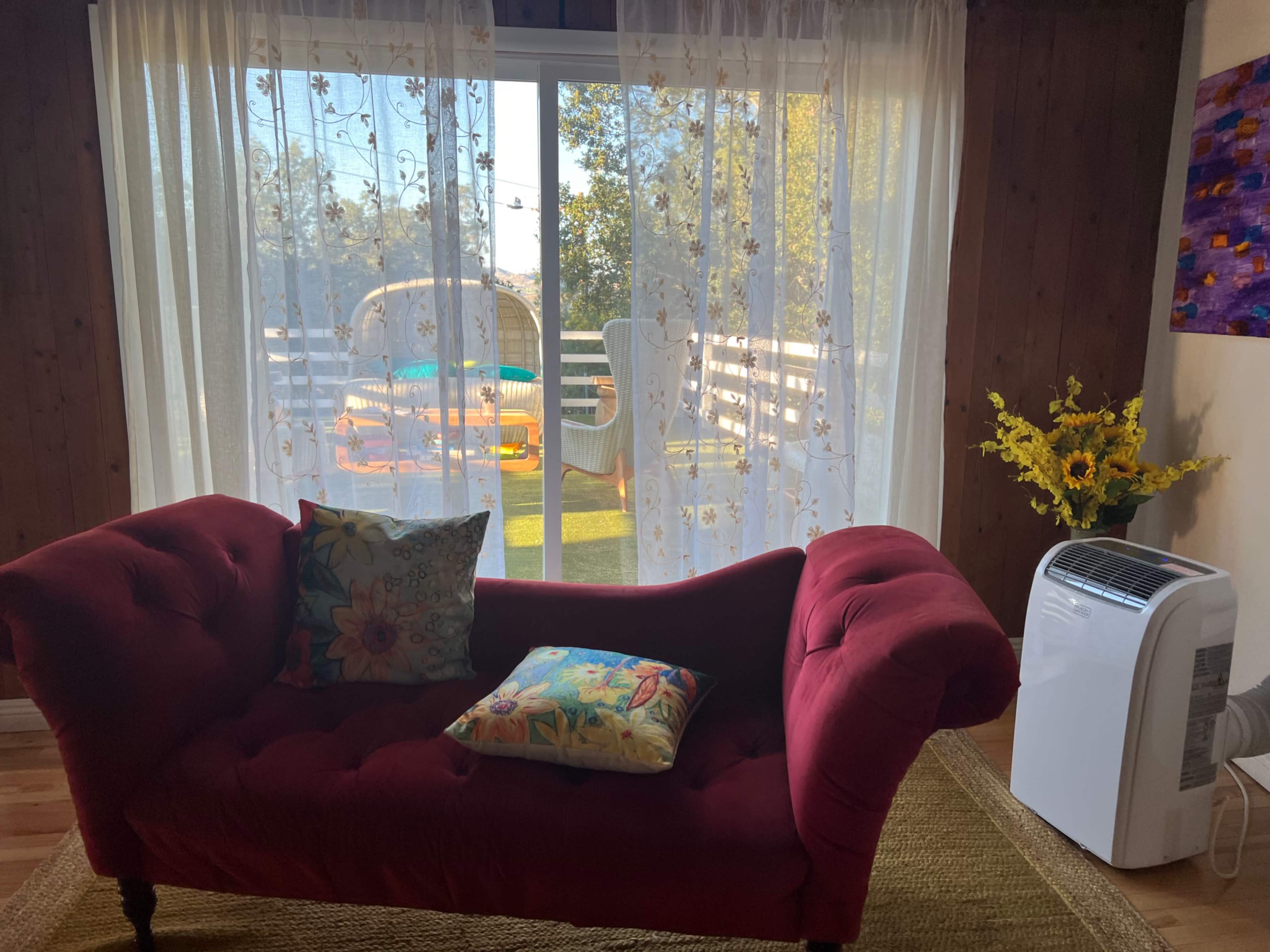 A red couch with decorative pillows sits in front of a sheer curtain, revealing a sunny outdoor space with chairs and plants.