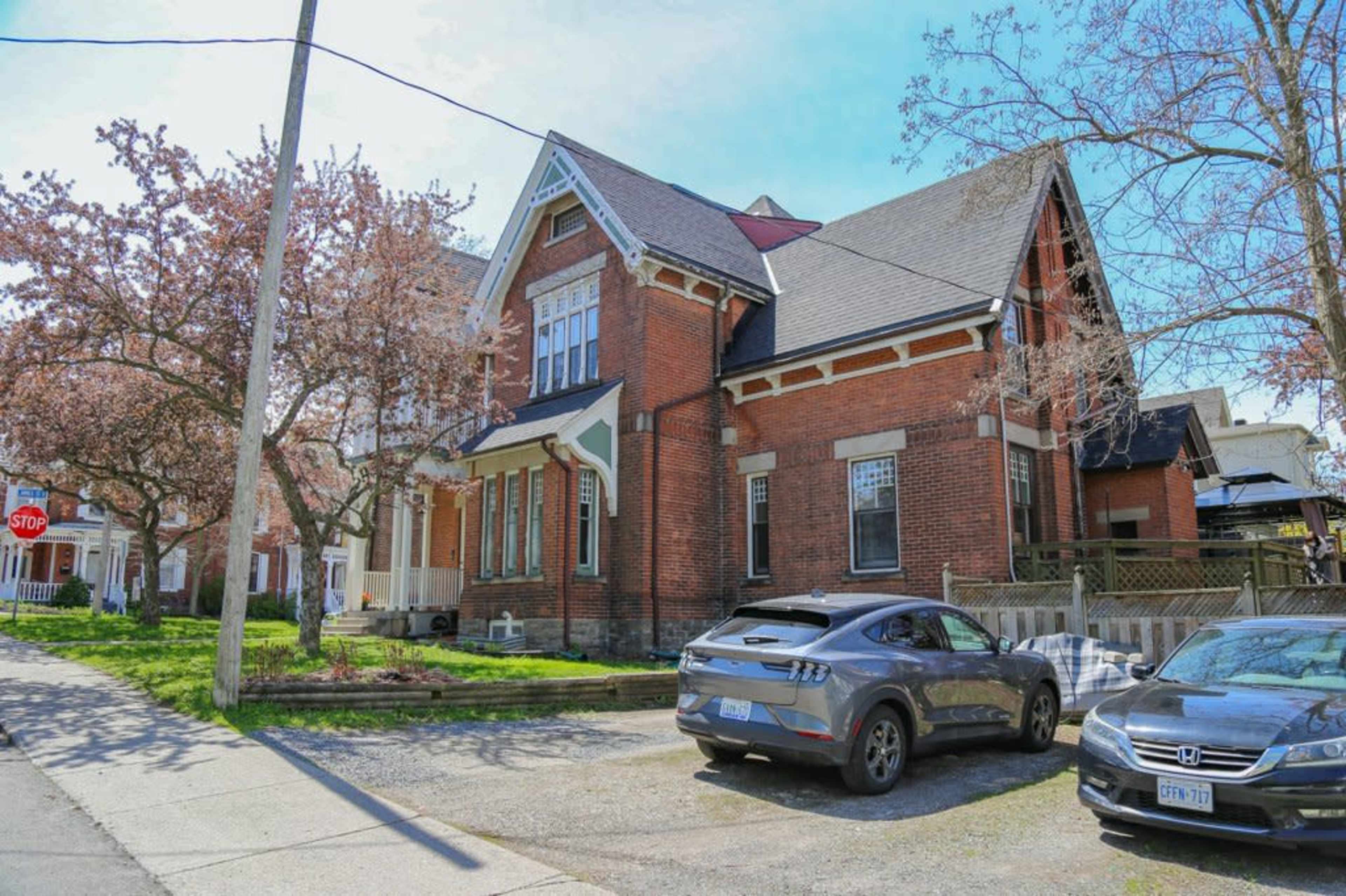 A red brick house with a steep roof is situated on a street lined with blooming trees, alongside parked cars in the driveway.