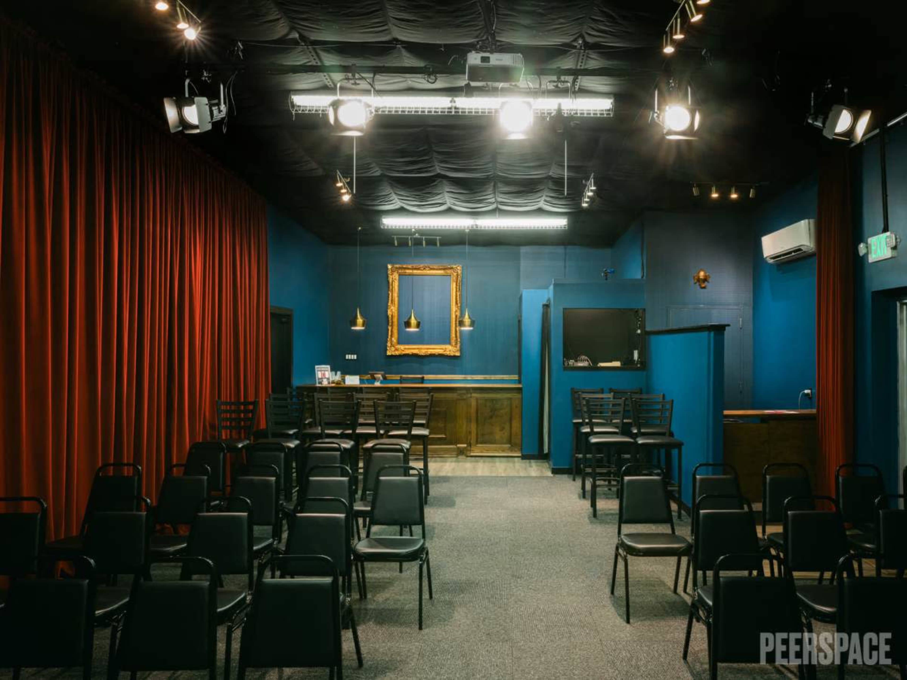 A small theater with black chairs arranged in rows, a blue wall, and a bar area featuring a framed gold mirror.