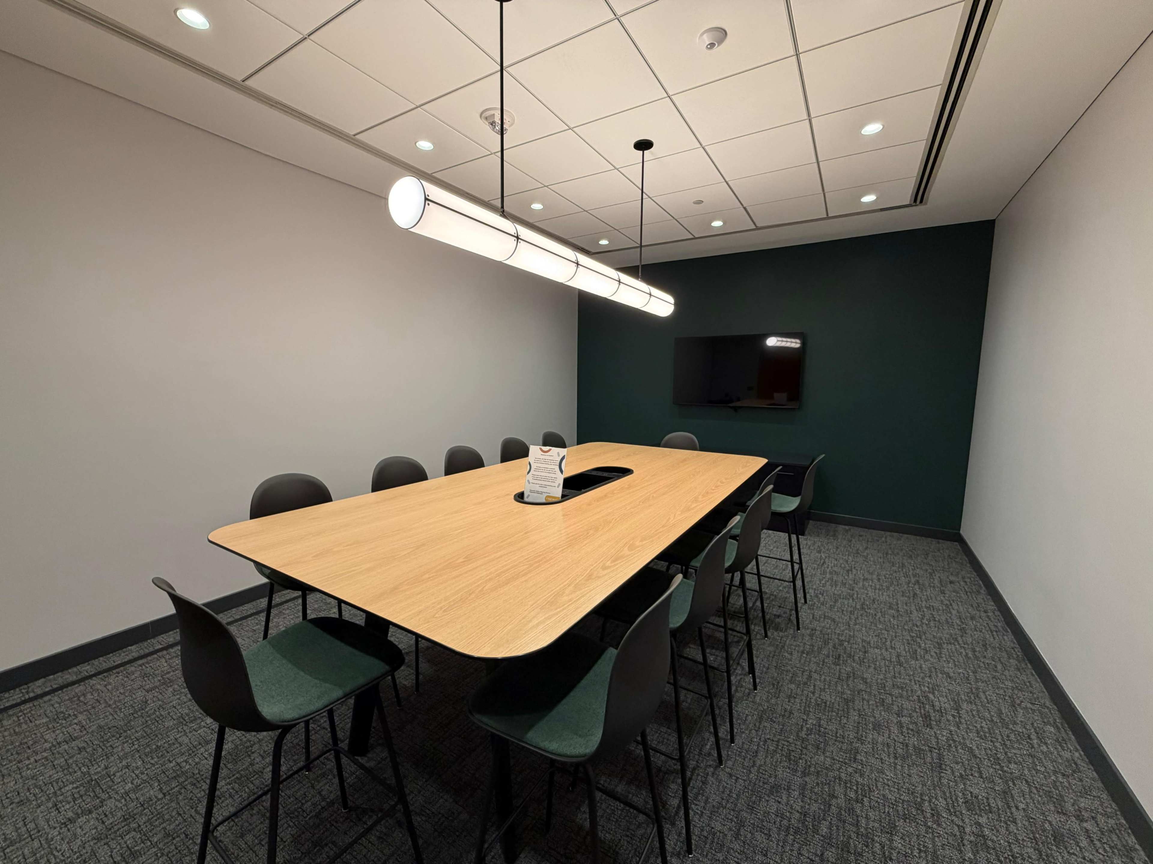A conference room features a large wooden table surrounded by black and green chairs, with a television mounted on the wall and a modern light fixture overhead.