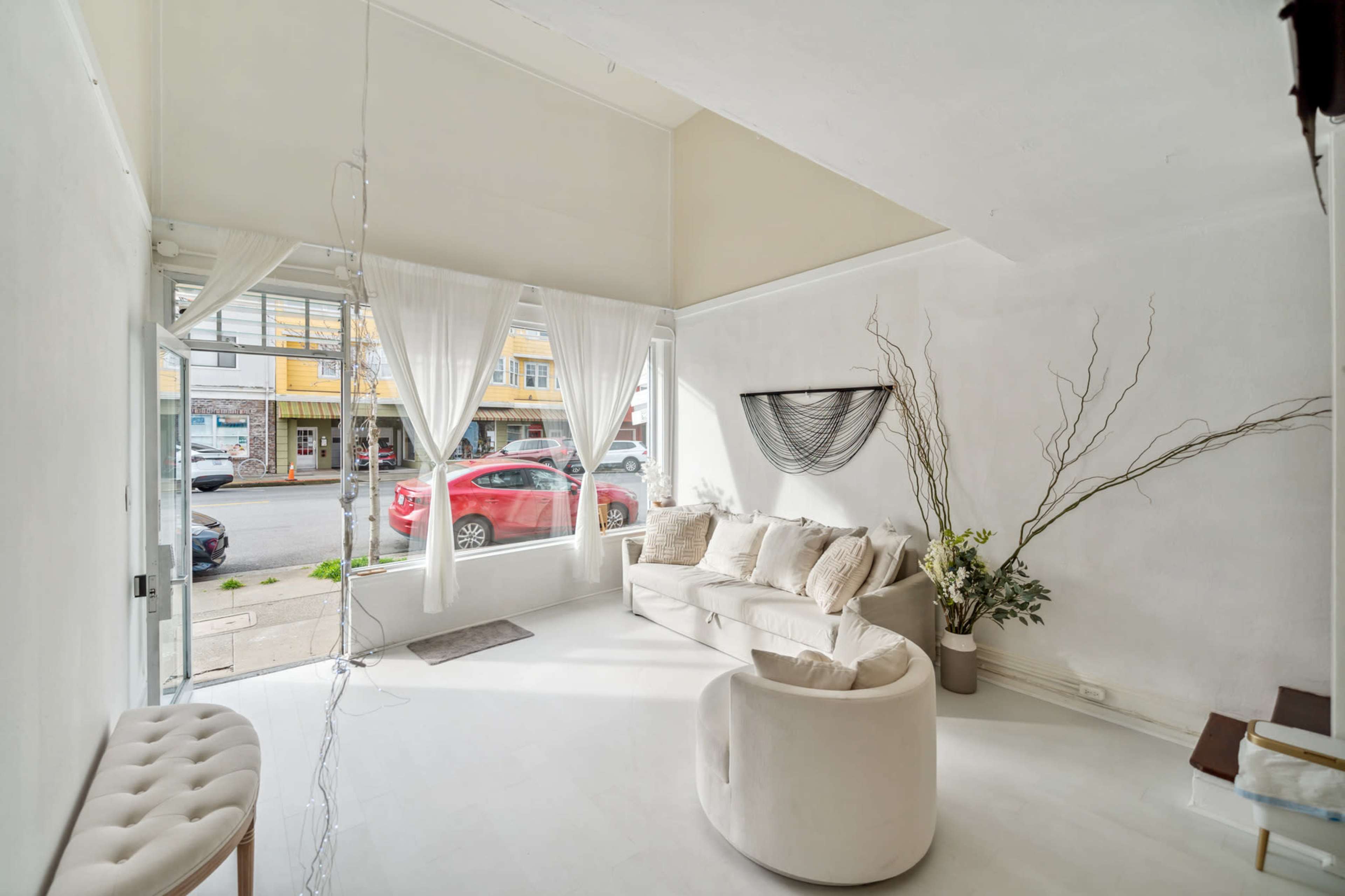 A bright, minimalistic interior of a modern space with a white couch, decorative branches, and large windows facing a street with parked cars.