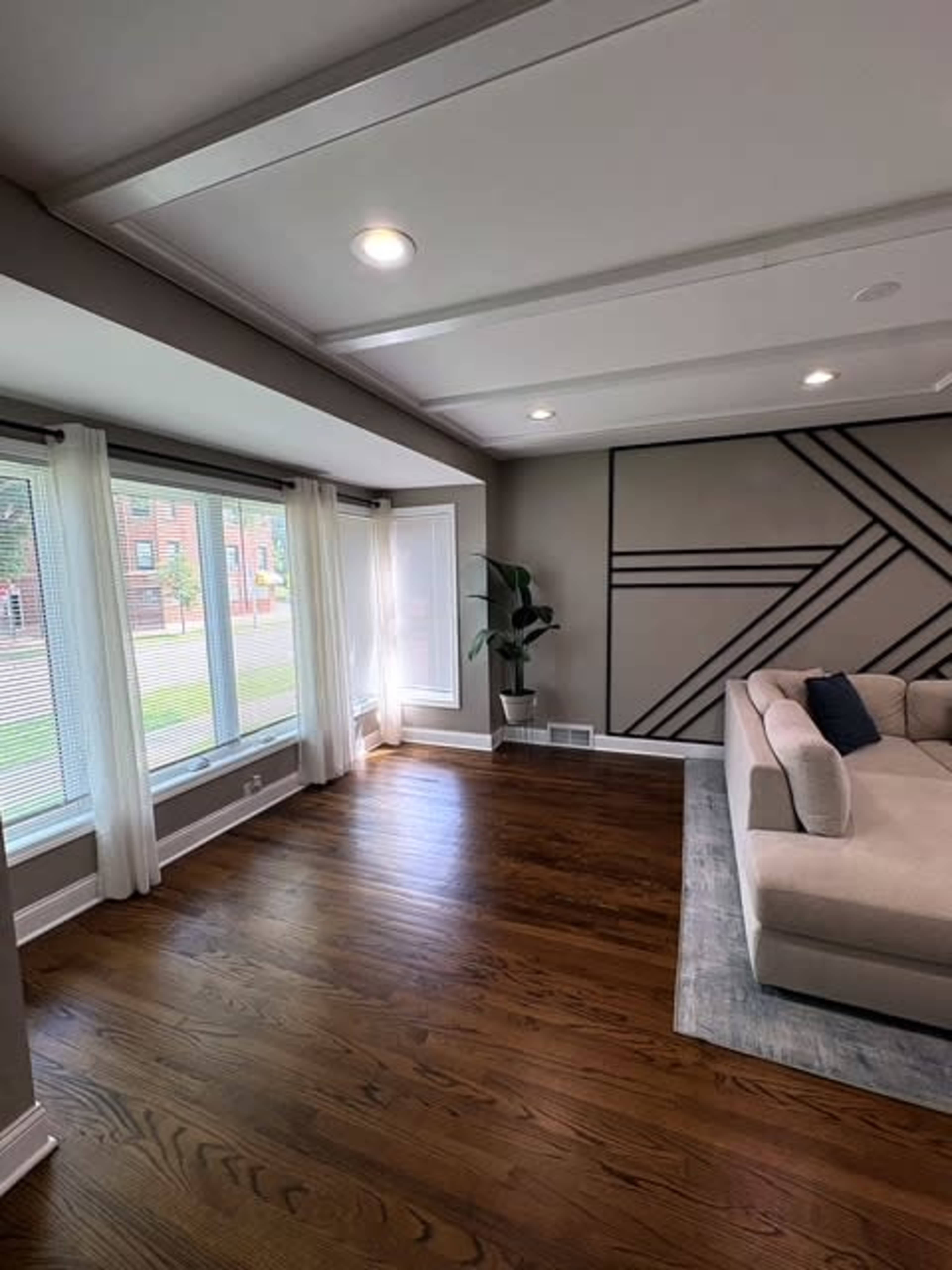 The image shows a bright, spacious living room with hardwood floors, large windows, and a sectional sofa.