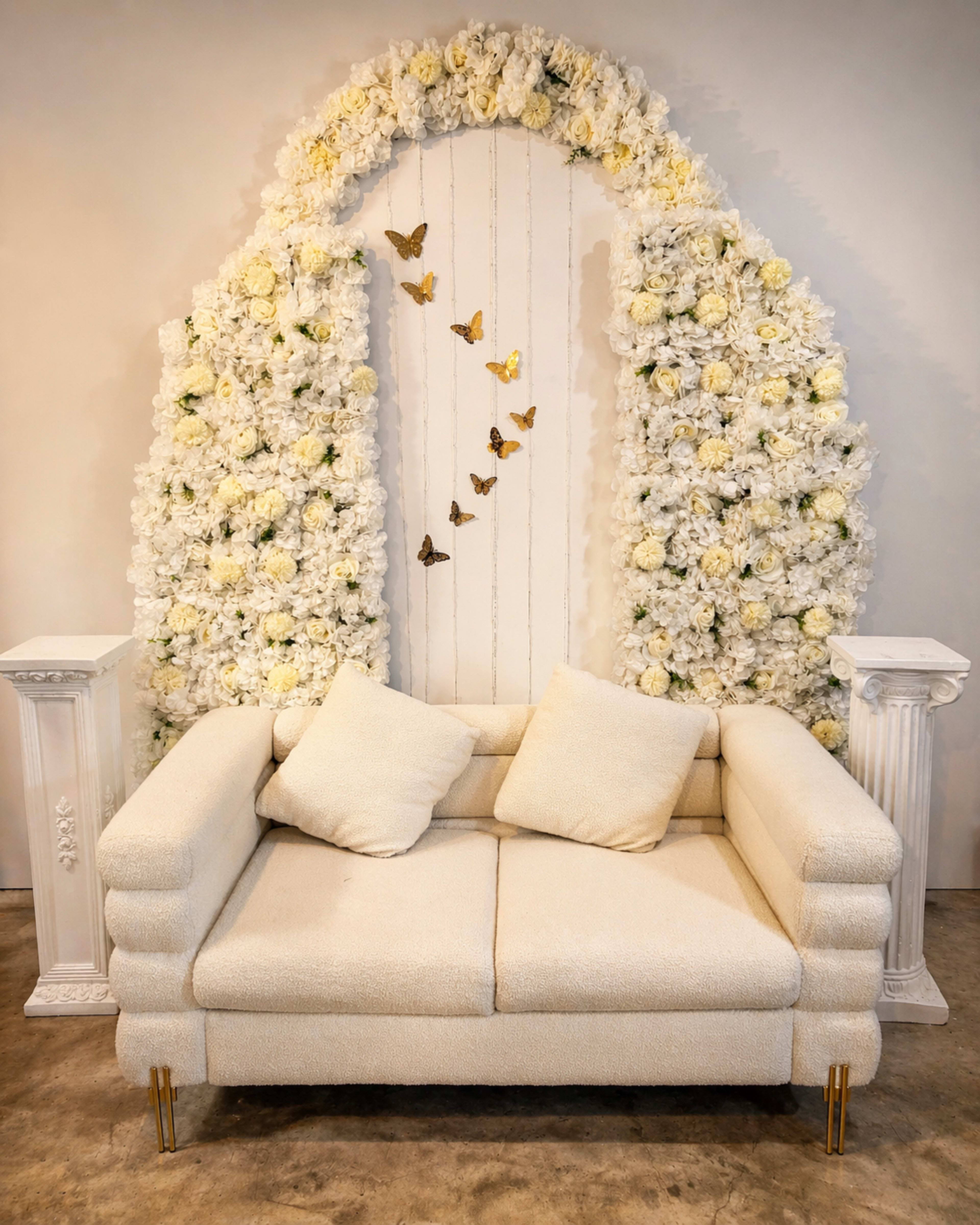 A white sofa with two cushions is positioned in front of a floral backdrop adorned with white flowers and decorative butterflies.