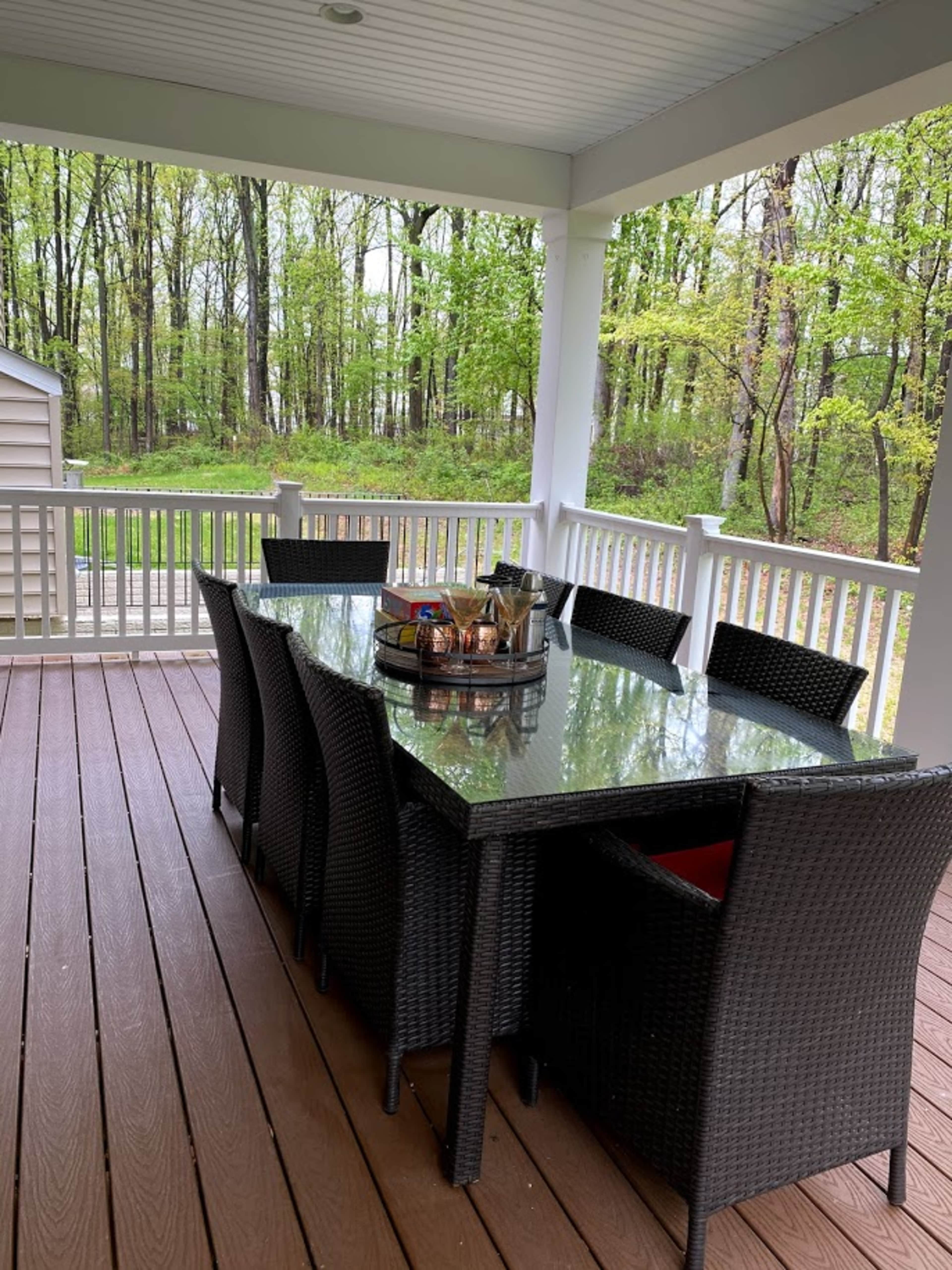 A rectangular dining table with eight chairs sits on a covered porch surrounded by trees.