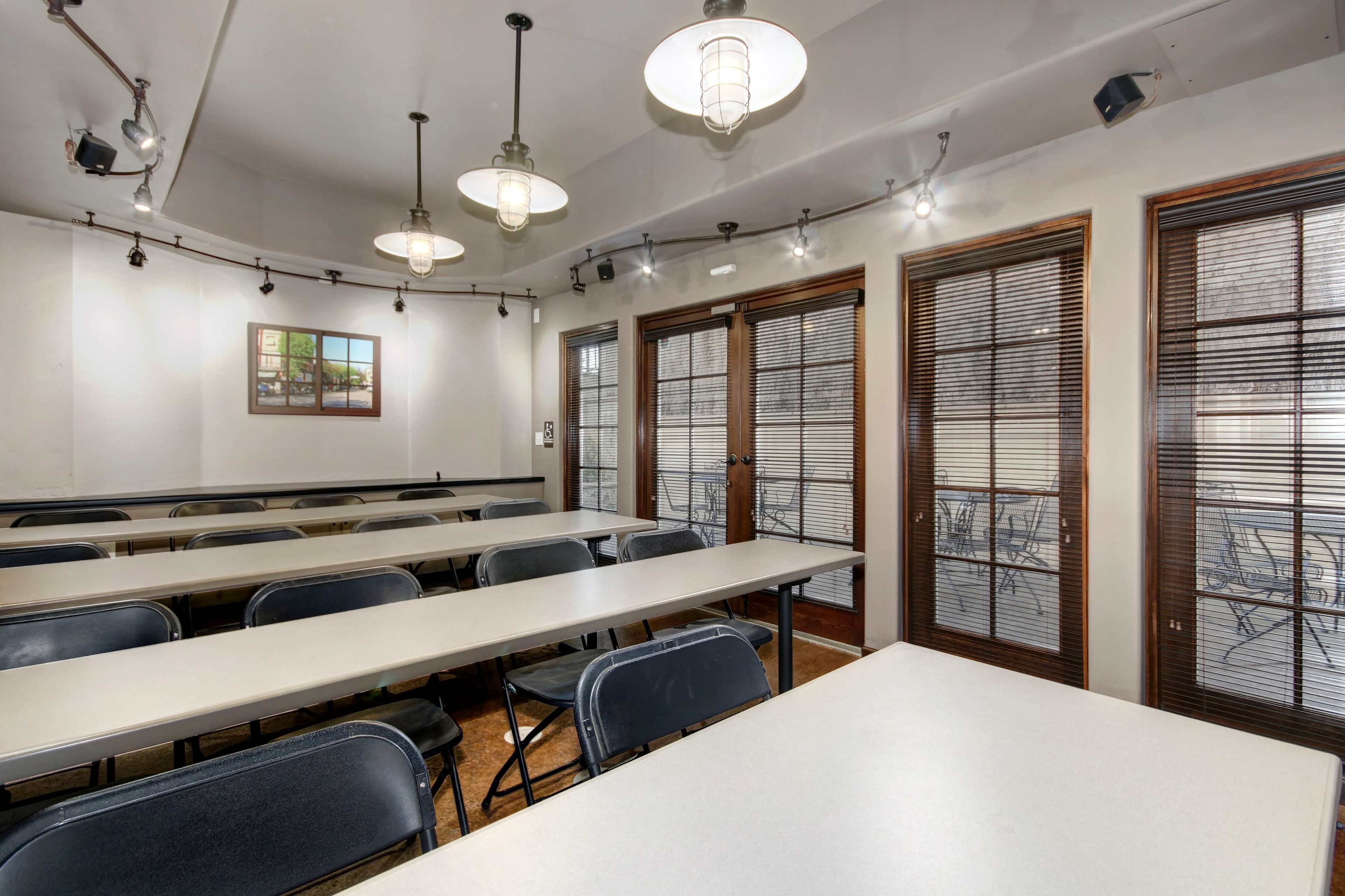 A classroom setting with several rows of tables and chairs, illuminated by overhead lights, and large windows featuring blinds.