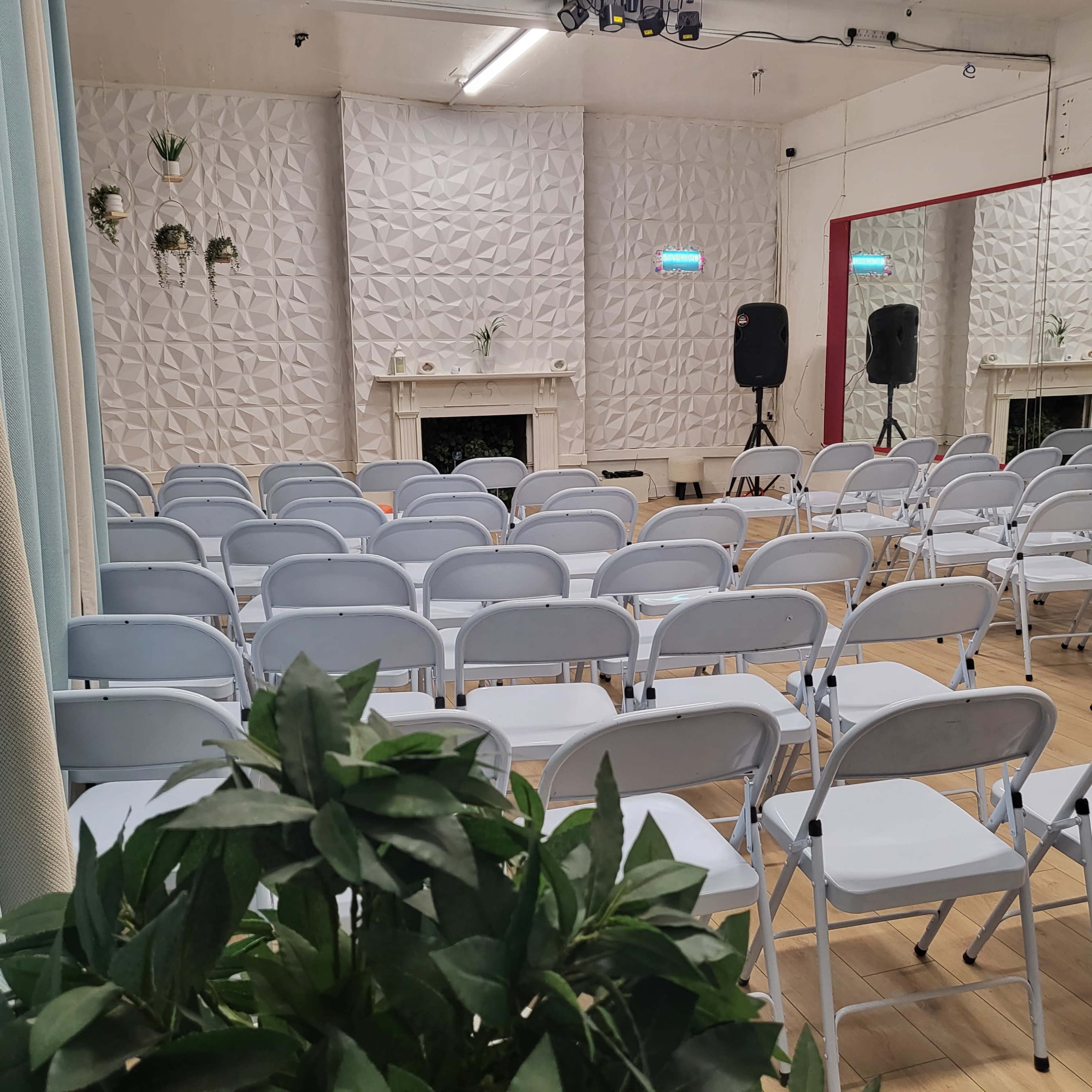 The image shows a brightly lit room set up with white folding chairs facing a fireplace, flanked by two speakers and a mirror reflecting the space.