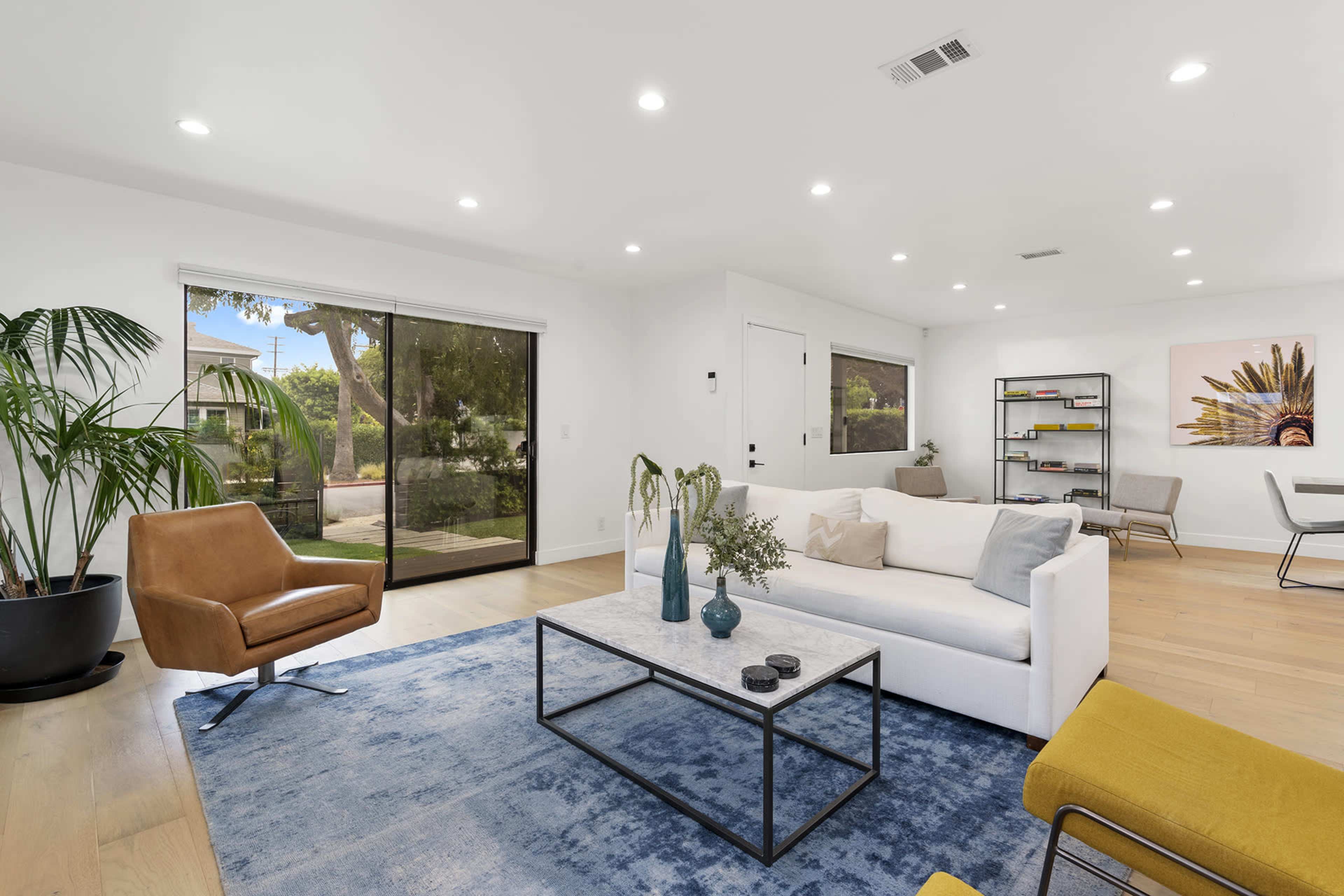 A modern living room features a white sofa, a glass coffee table, and a brown armchair, with large windows overlooking a garden area.