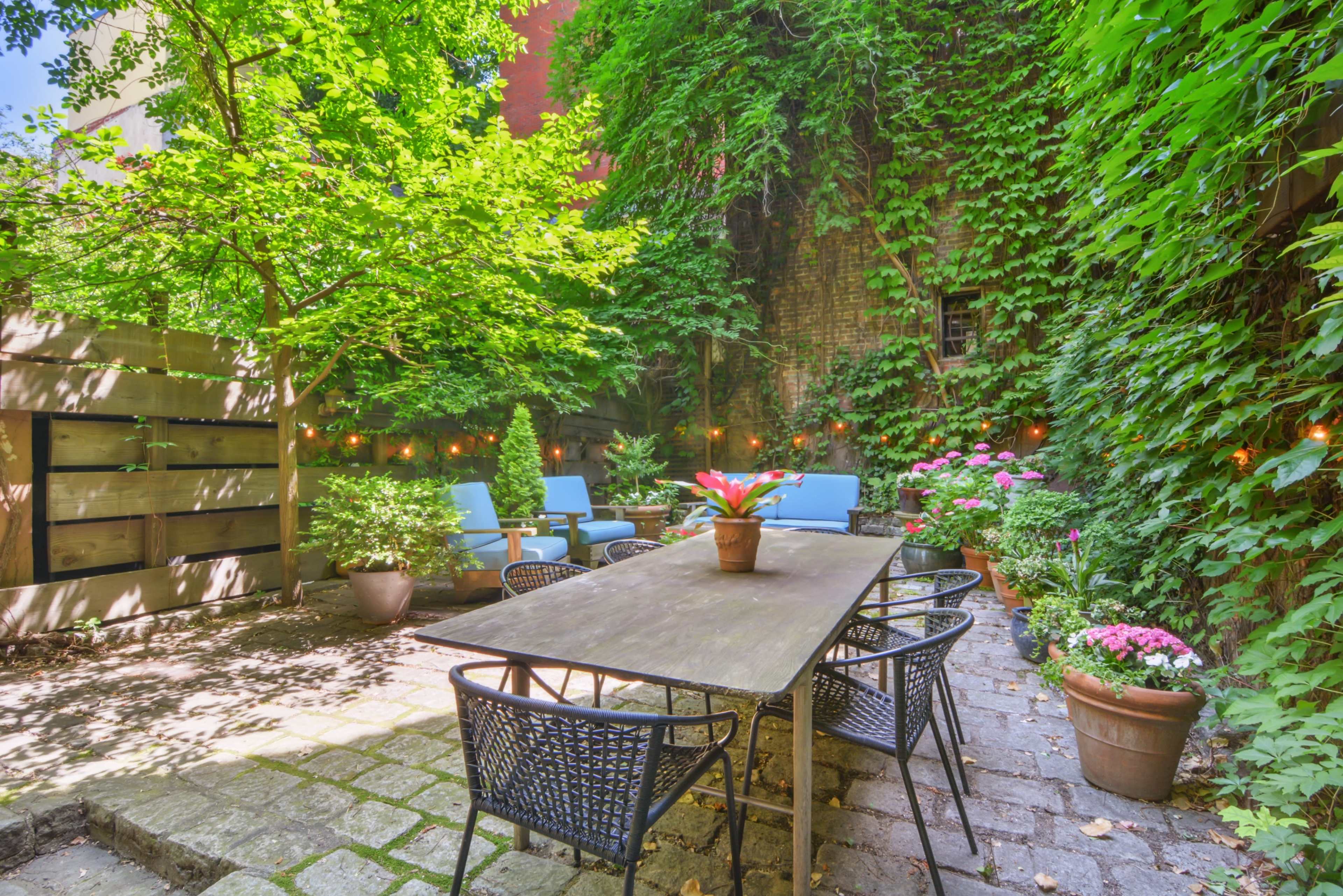 A shaded outdoor patio features a wooden table surrounded by black chairs, with potted plants and decorative lights in a green, leafy setting.